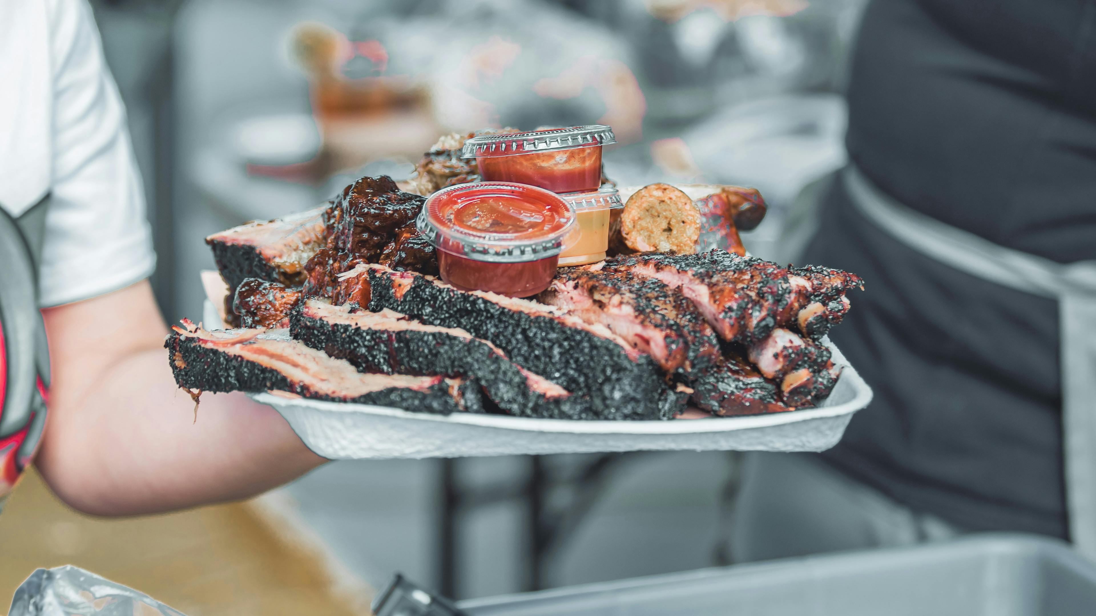A person is holding a white paper platter piled high with smoked and grilled meats, including sliced brisket and ribs, with small containers of sauce on top.