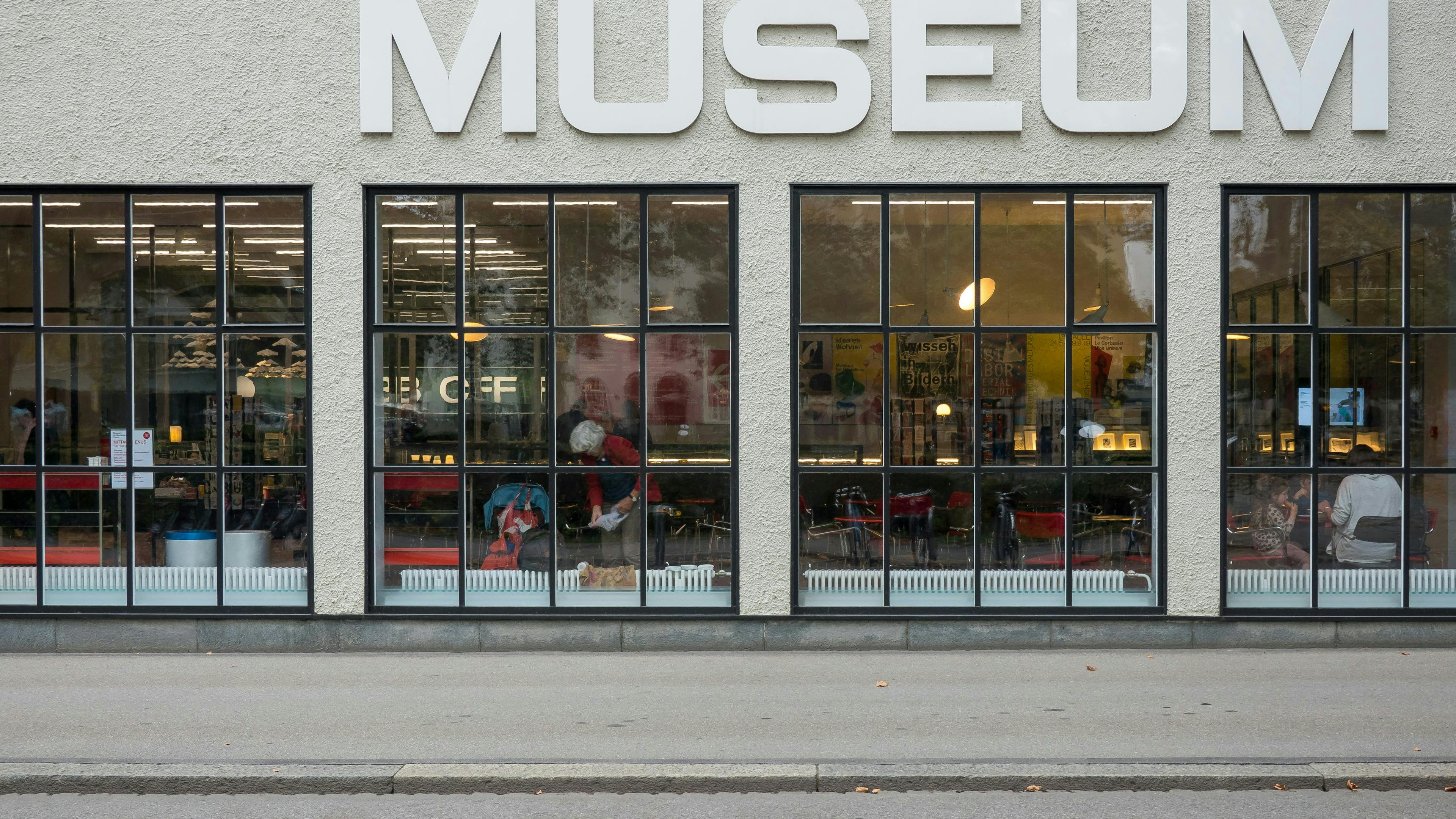 The gray concrete exterior of a museum with large windows and a modern looking interior