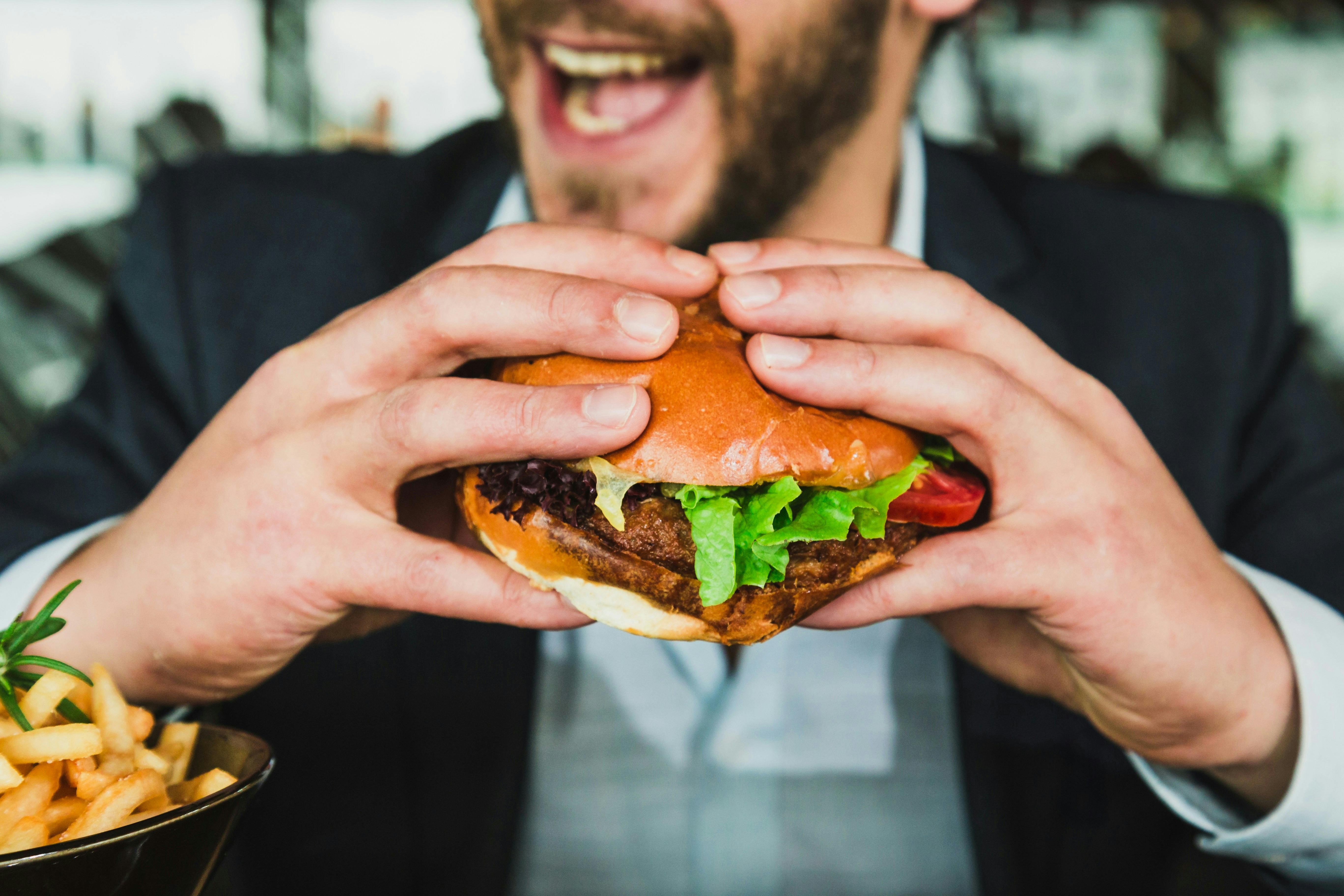 A close-up shot of a smiling man in a suit holding a large hamburger with both hands. A bowl of french fries is visible to the left.