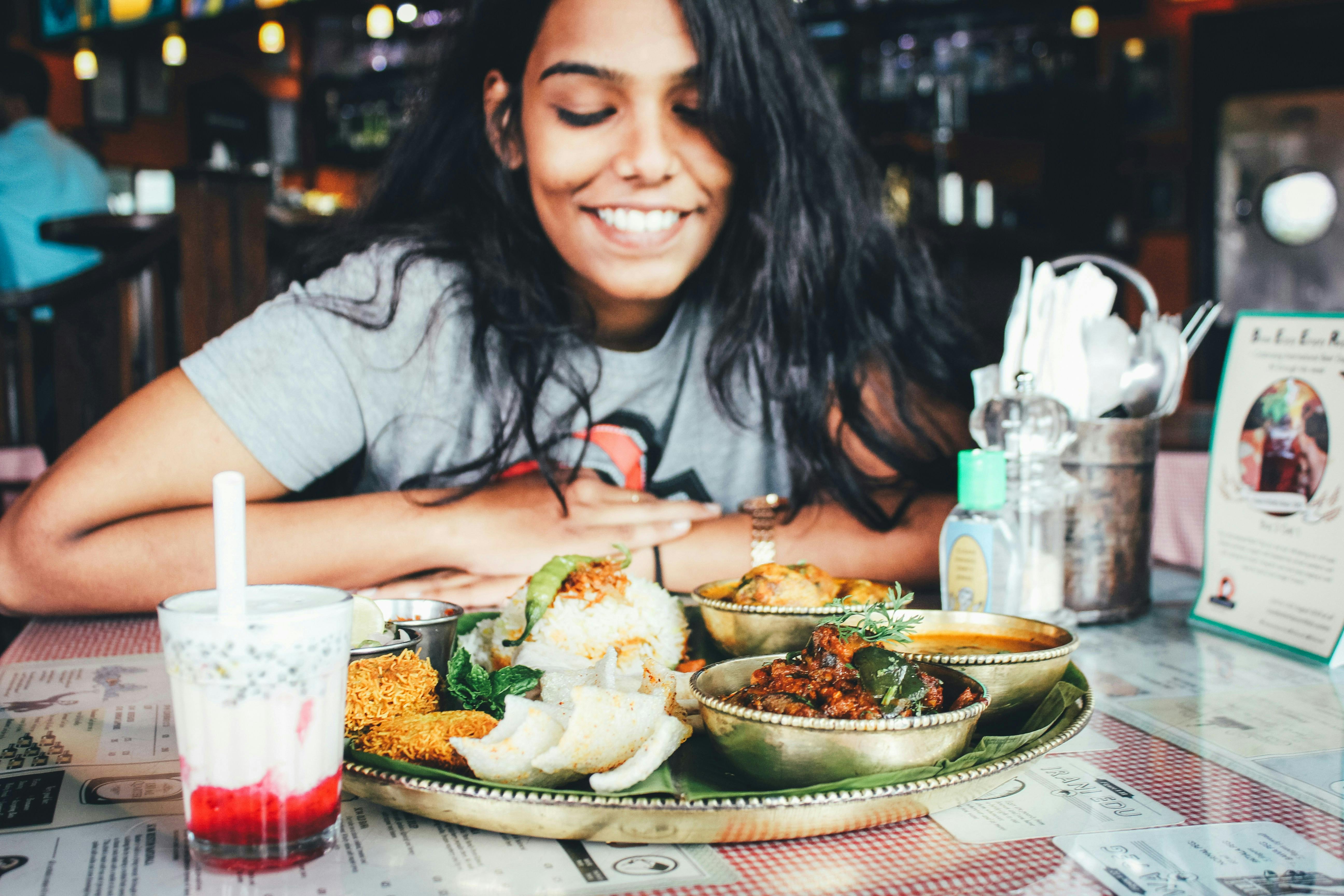 A smiling woman with long dark hair is looking down at a traditional Indian thali platter with multiple bowls of food on a table. A glass of a red and white layered drink is in the foreground.