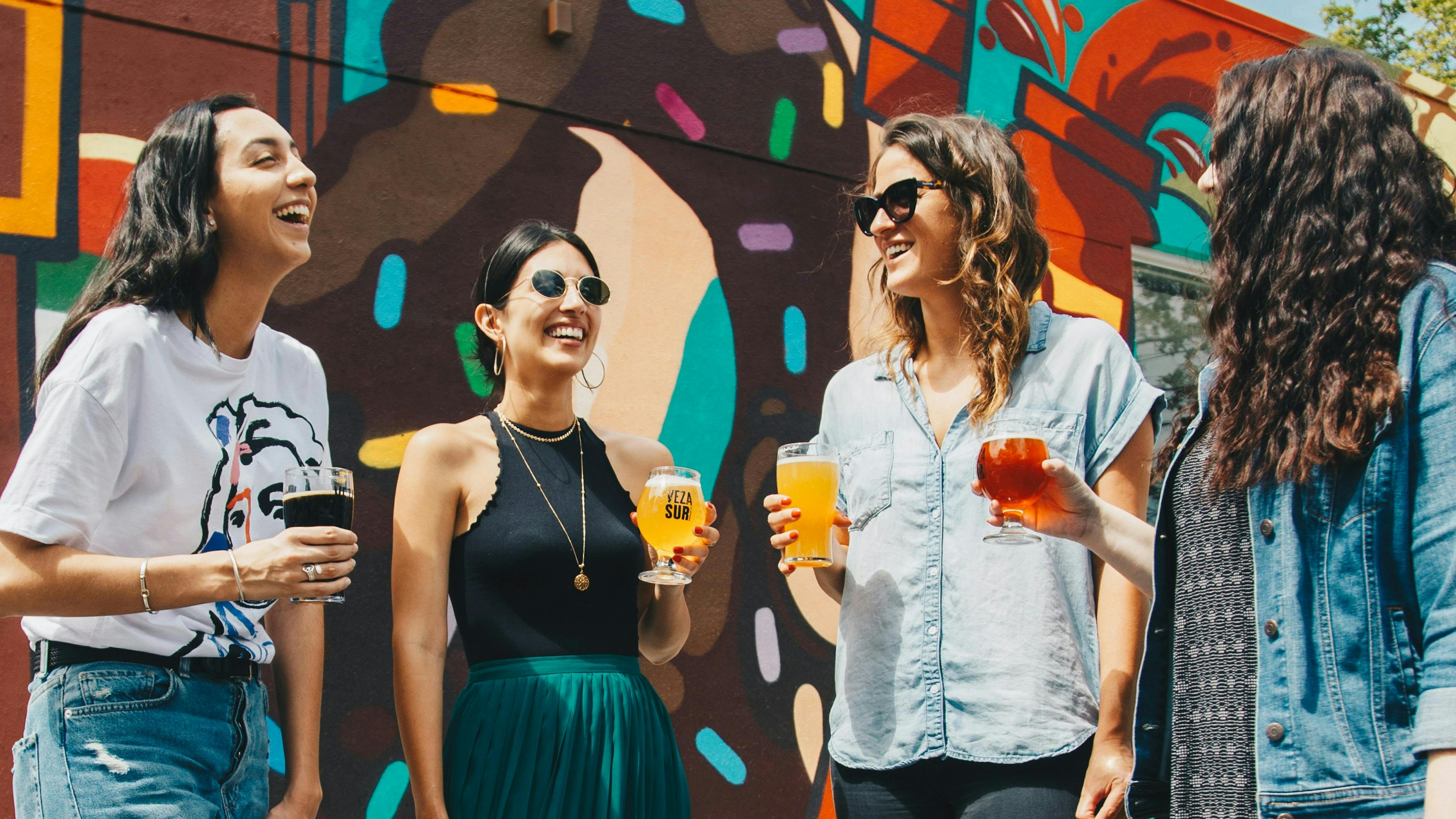 Four women are standing outside, laughing and holding glasses of beer. They are standing in front of a colorful mural.