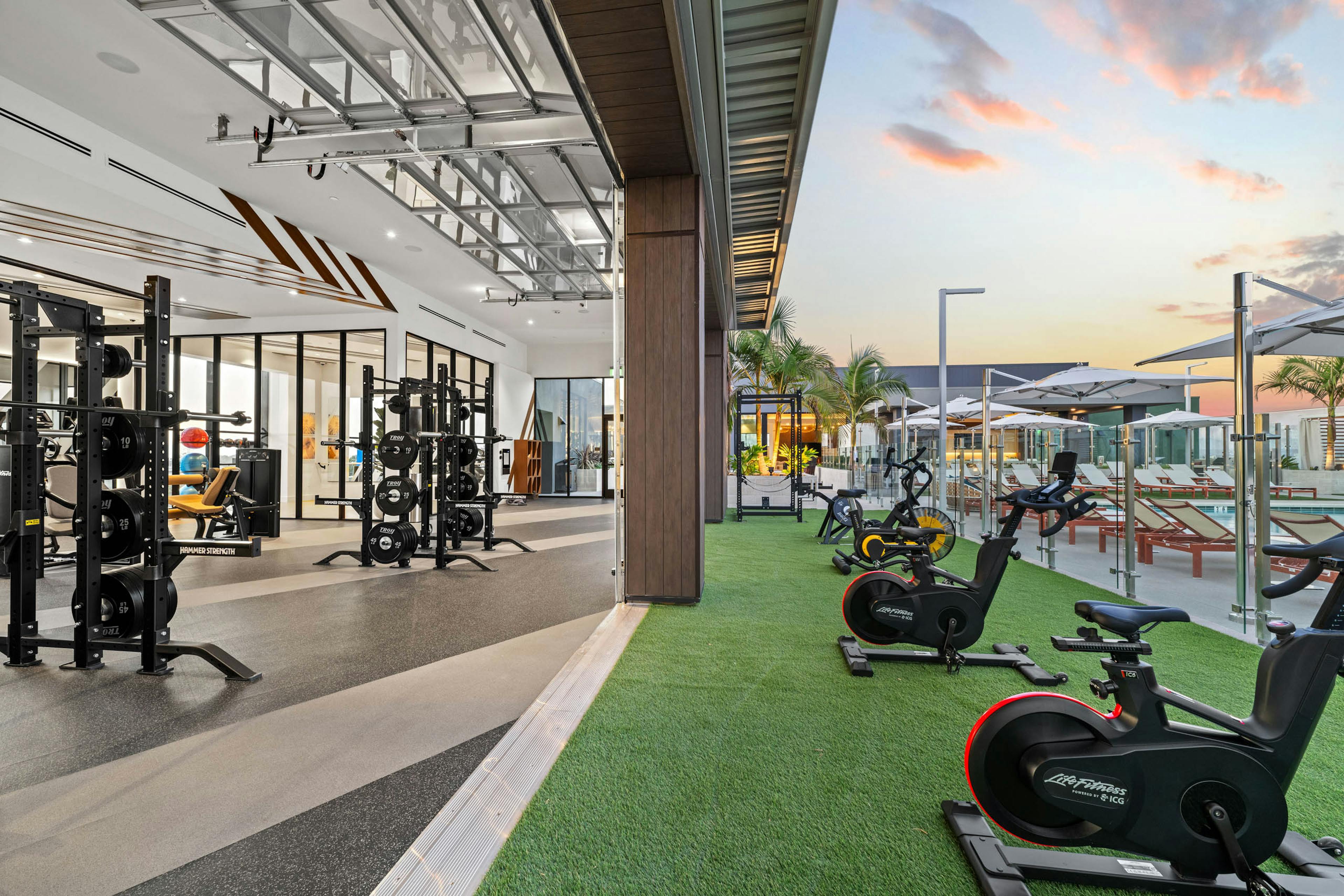 Indoor outdoor fitness at AMLI Aero apartments with weight racks inside and stationary bikes on turf overlooking rooftop pool deck.