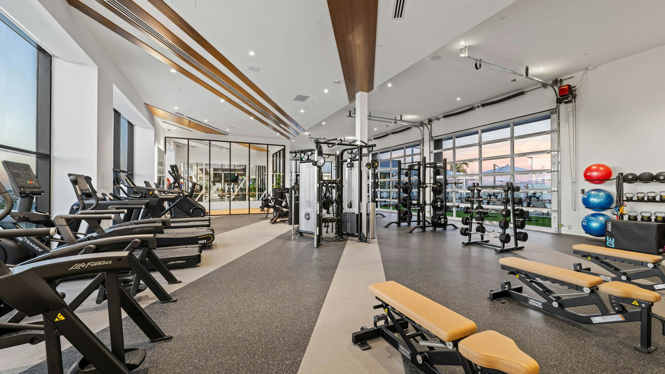 Fitness center at AMLI Aero apartments with treadmills, weight machines, benches, and floor-to-ceiling windows with bright lighting.