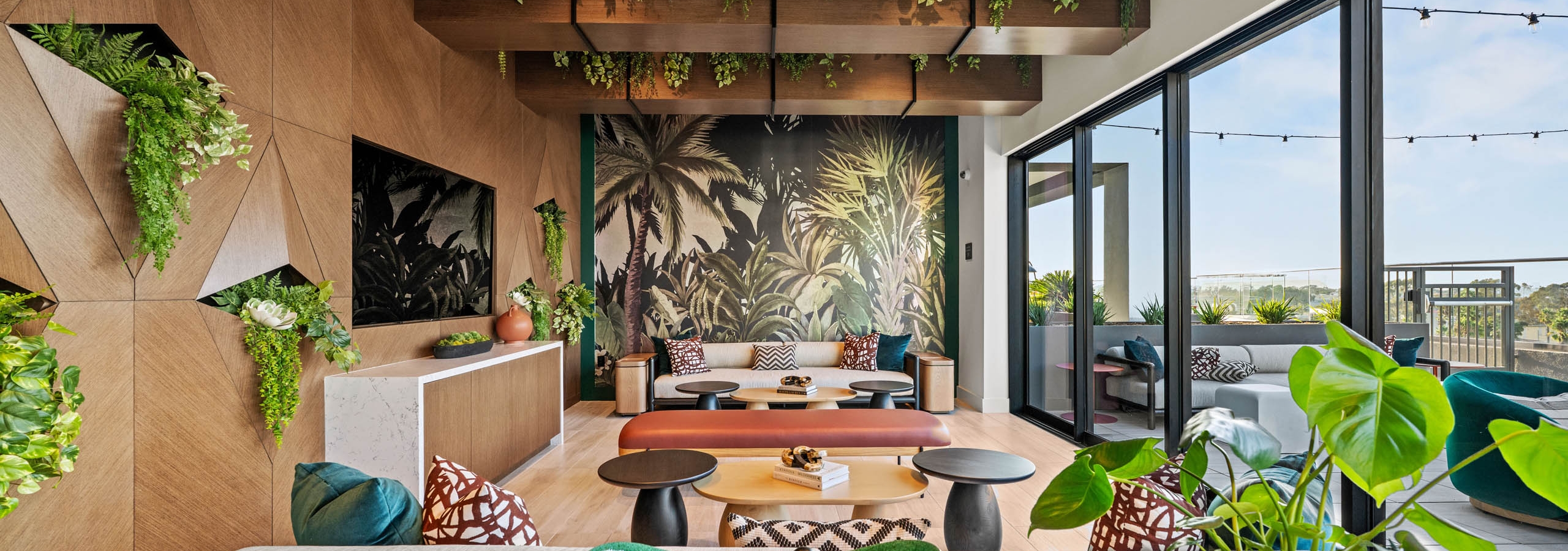 Sunset lounge at AMLI Aero apartments with tropical mural, ceiling greenery, mounted TV modern seating, and glass doors opening to terrace.