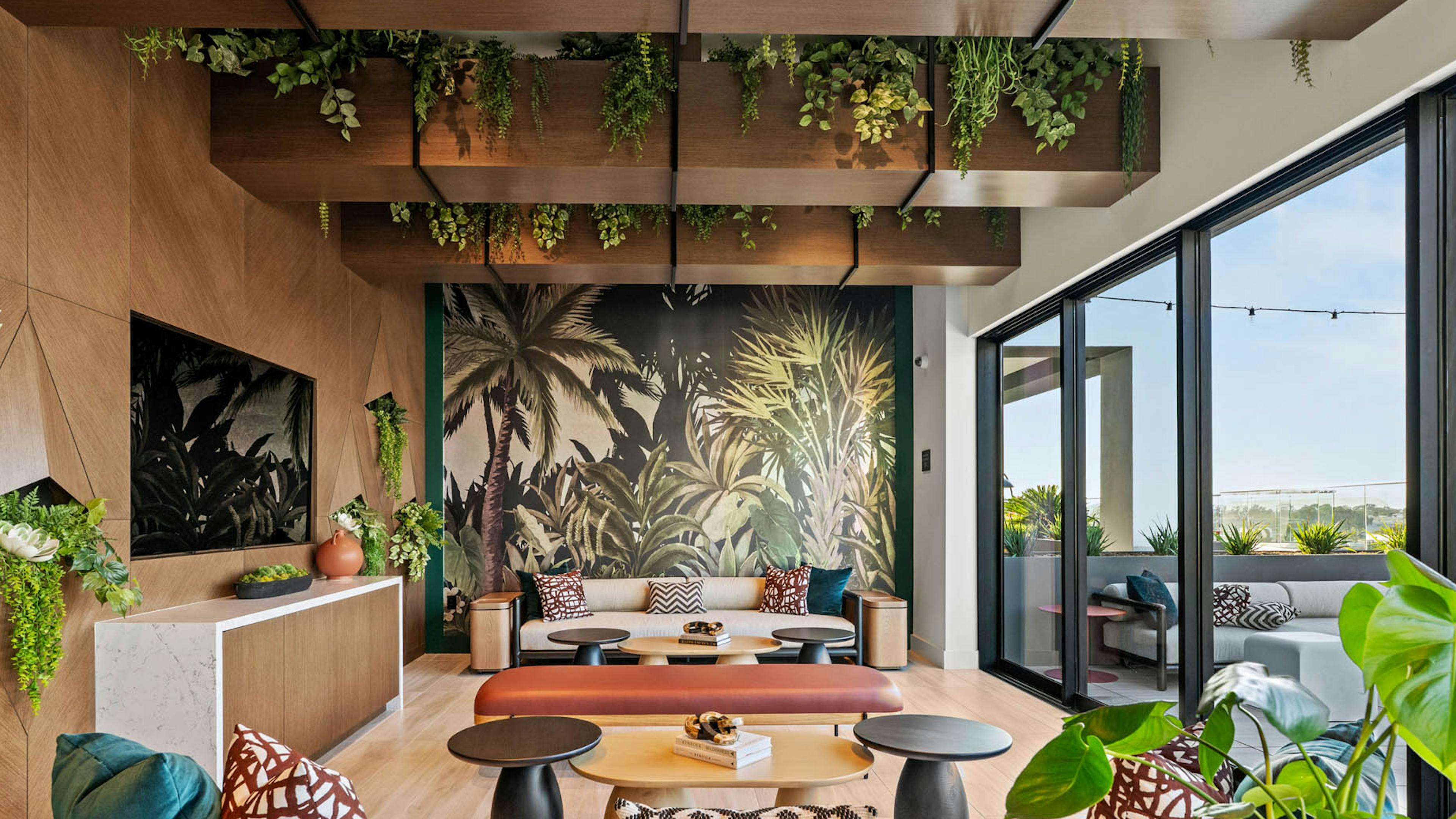 Sunset lounge at AMLI Aero apartments with tropical mural, ceiling greenery, mounted TV modern seating, and glass doors opening to terrace.