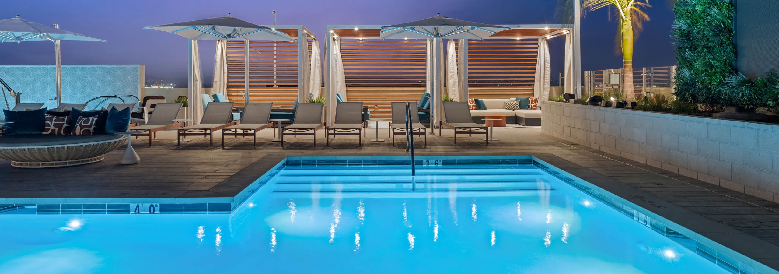 Cabanas with wood slat accents at AMLI Aero apartments rooftop pool with lounge chairs, umbrellas, palm trees, and under evening sky.