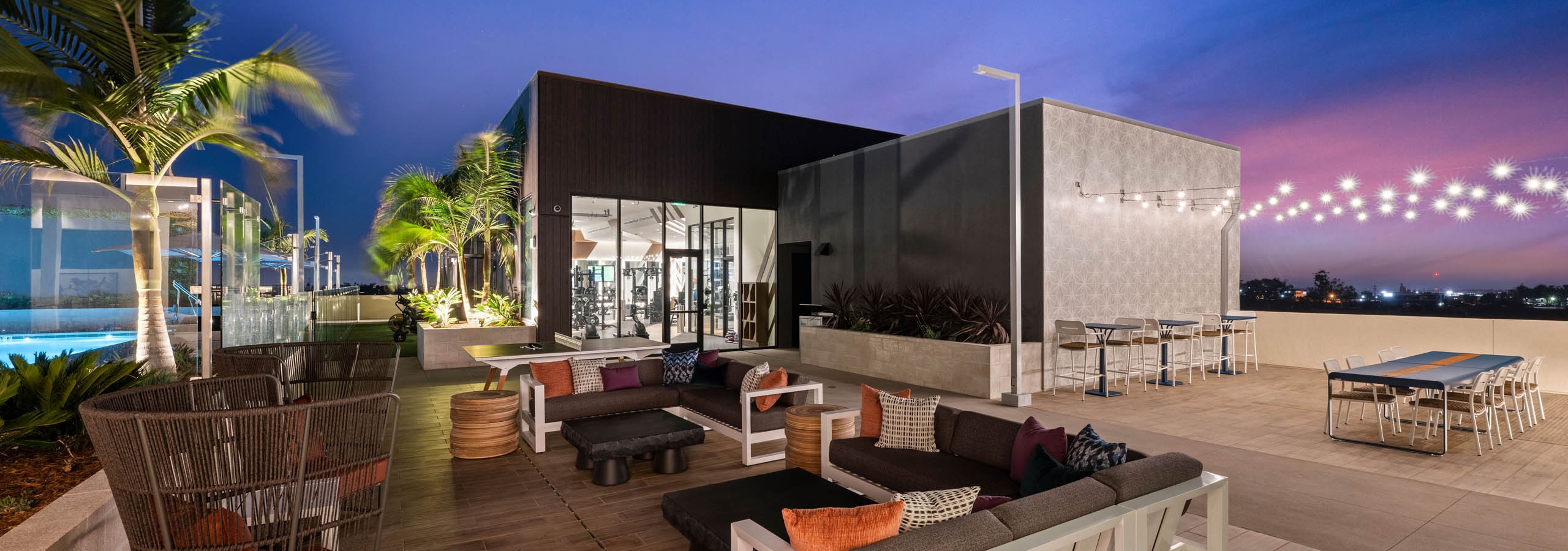 Rooftop terrace at AMLI Aero apartments with cushioned lounge seating, palm trees, string lights, and dining tables at twilight.