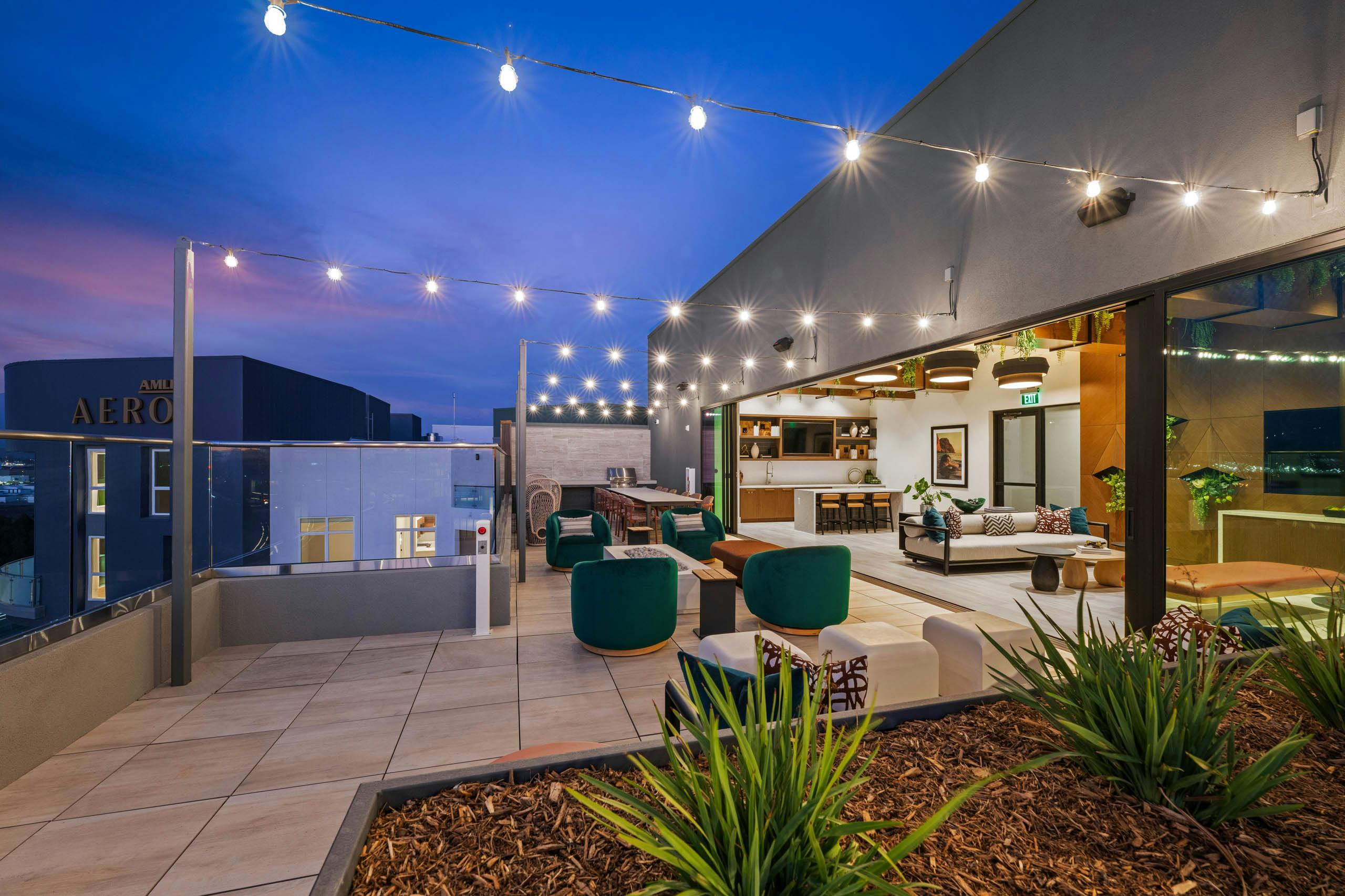 Evening view of rooftop lounge at AMLI Aero apartments with string lights, cushioned seating, and indoor-outdoor clubroom with kitchen.