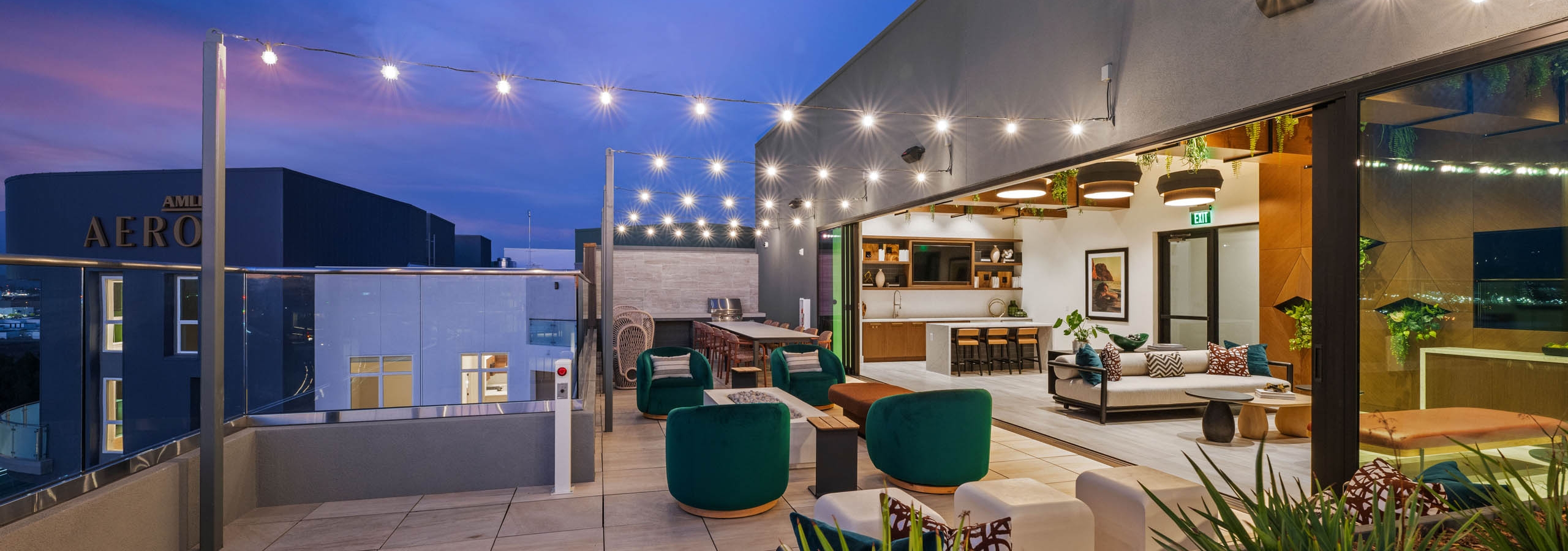 Evening view of rooftop lounge at AMLI Aero apartments with string lights, cushioned seating, and indoor-outdoor clubroom with kitchen.