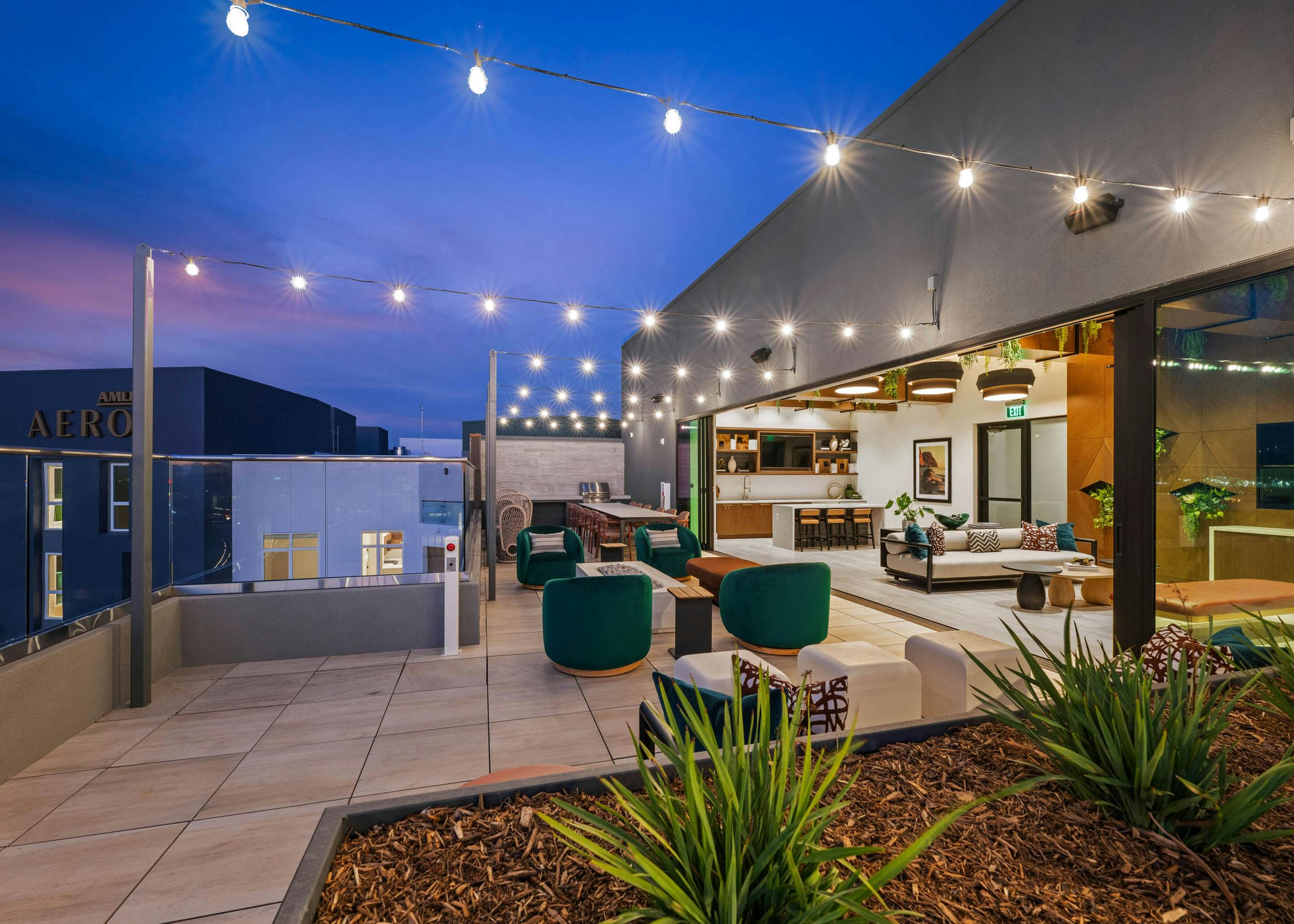 Evening view of rooftop lounge at AMLI Aero apartments with string lights, cushioned seating, and indoor-outdoor clubroom with kitchen.