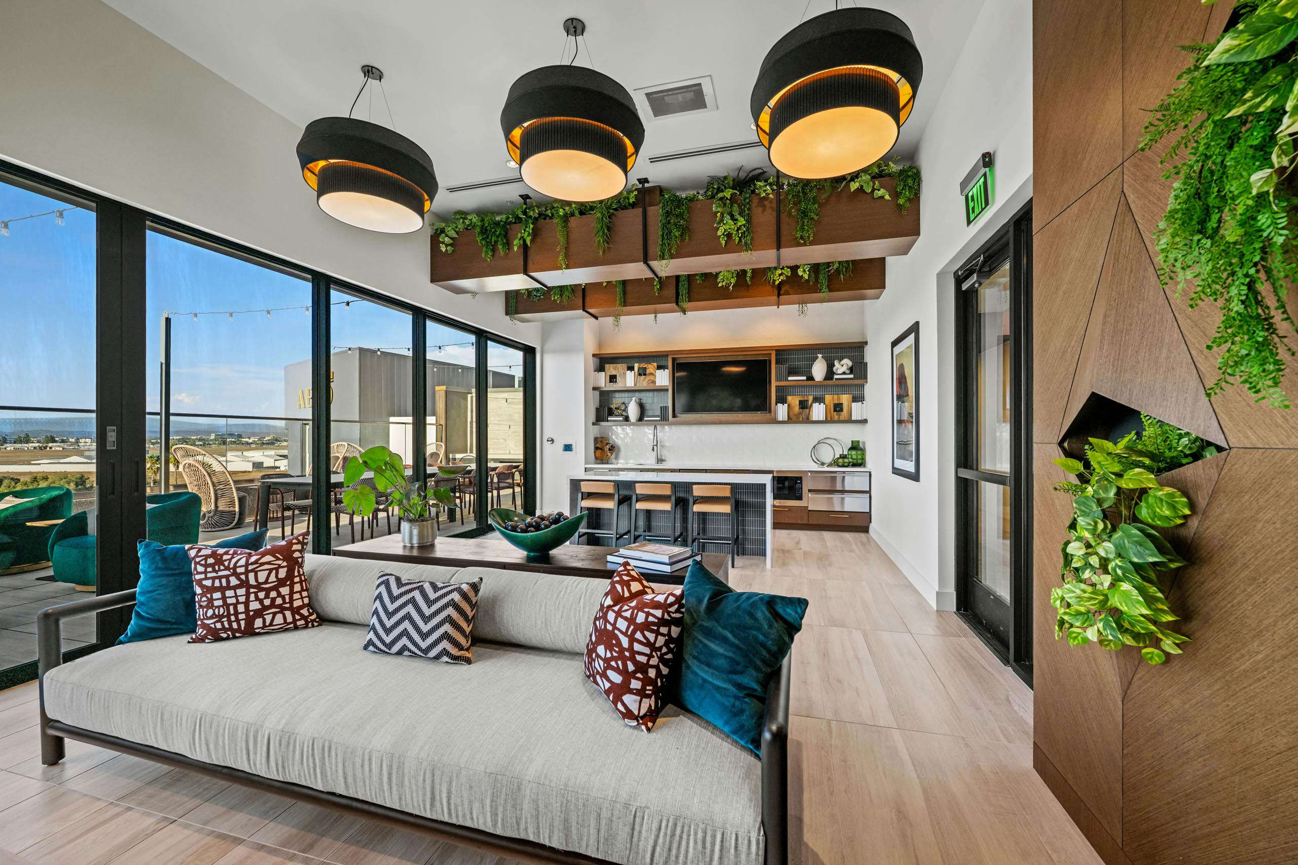 Interior daytime view of AMLI Aero apartments Sunset rooftop lounge with sofa, hanging lights, greenery, and kitchen with island seating.