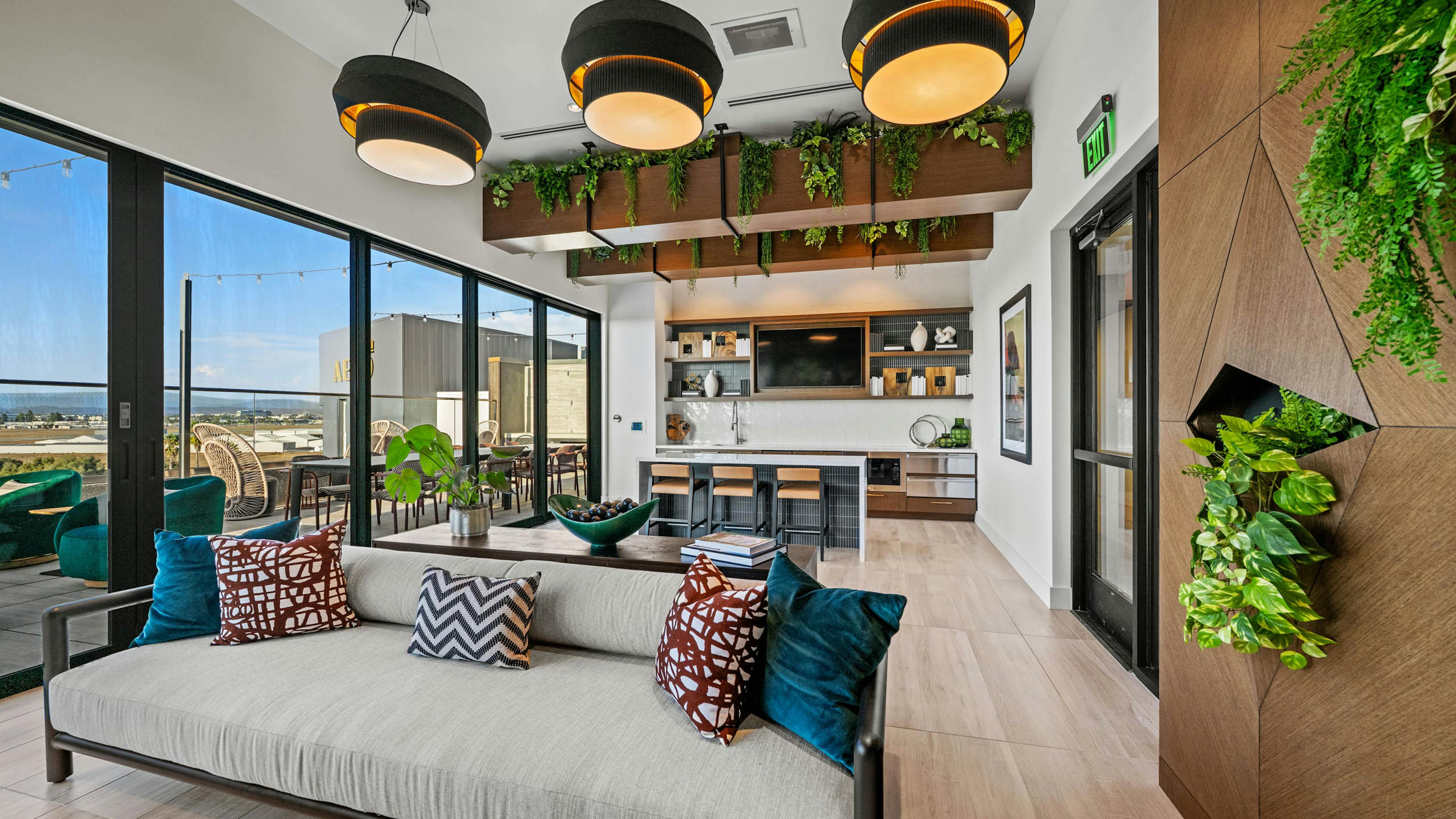 Interior daytime view of AMLI Aero apartments Sunset rooftop lounge with sofa, hanging lights, greenery, and kitchen with island seating.