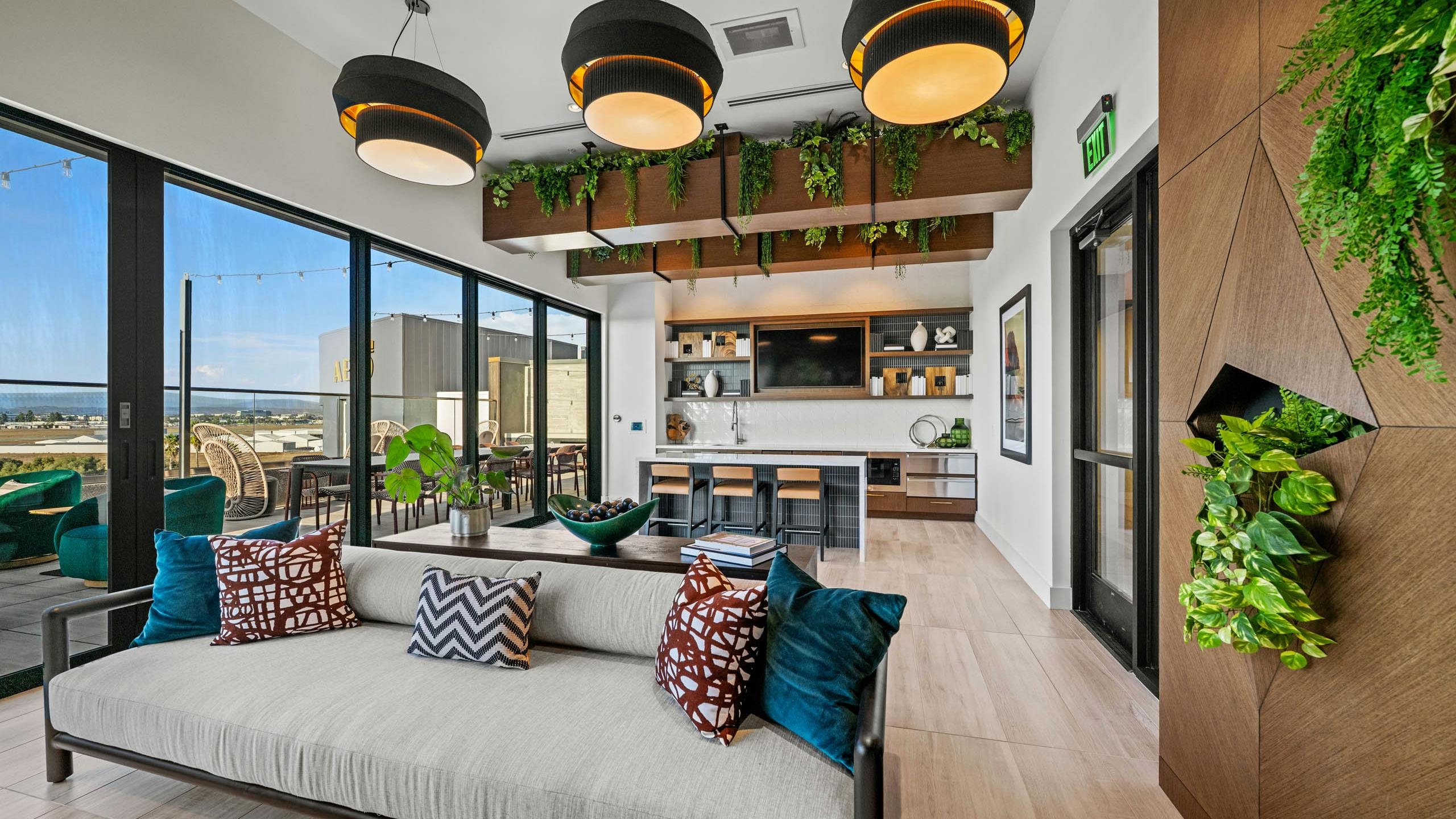 Interior daytime view of AMLI Aero apartments Sunset rooftop lounge with sofa, hanging lights, greenery, and kitchen with island seating.
