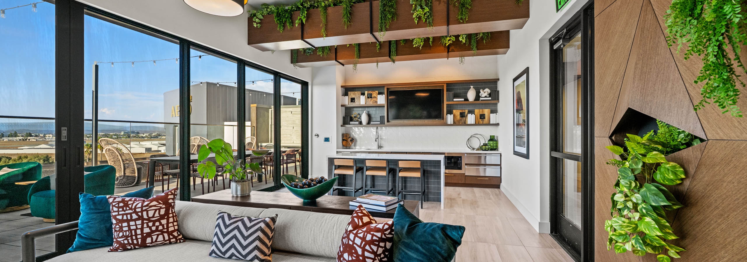 Interior daytime view of AMLI Aero apartments Sunset rooftop lounge with sofa, hanging lights, greenery, and kitchen with island seating.