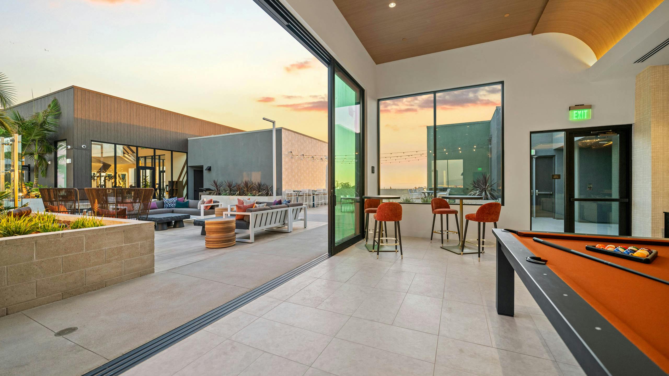 Rooftop clubroom at AMLI Aero apartments with pool table, tall windows, bar seating, and doors opening to rooftop terrace at sunset.