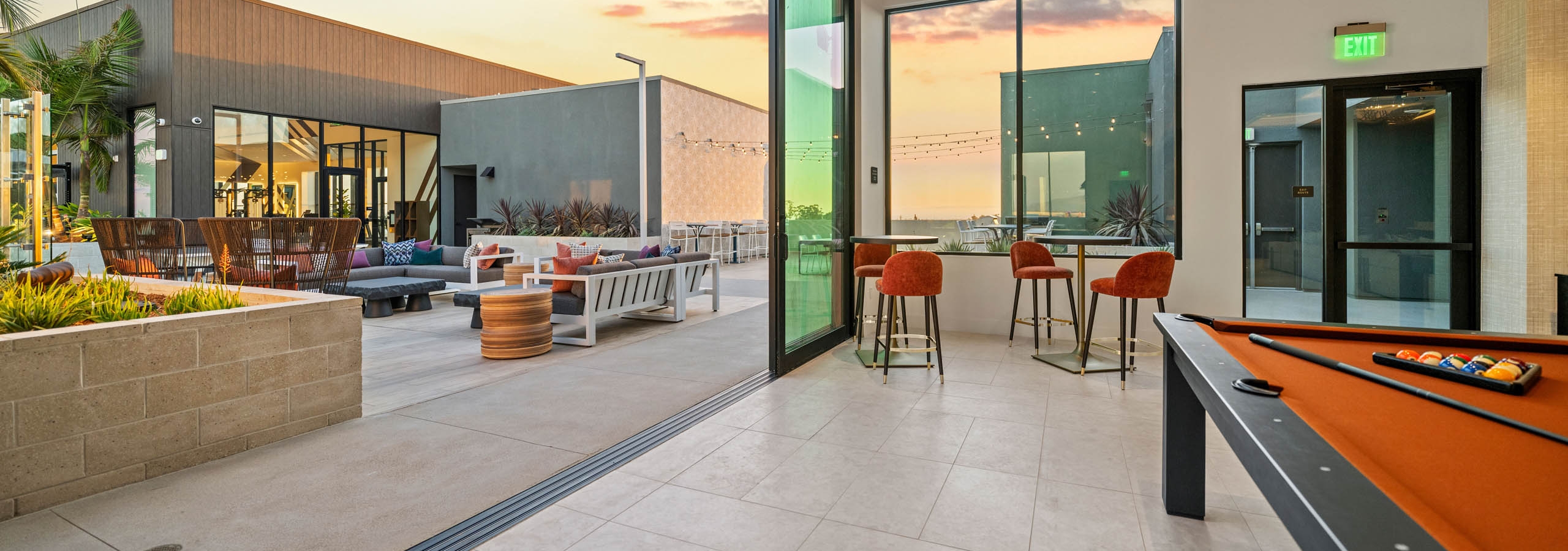 Rooftop clubroom at AMLI Aero apartments with pool table, tall windows, bar seating, and doors opening to rooftop terrace at sunset.