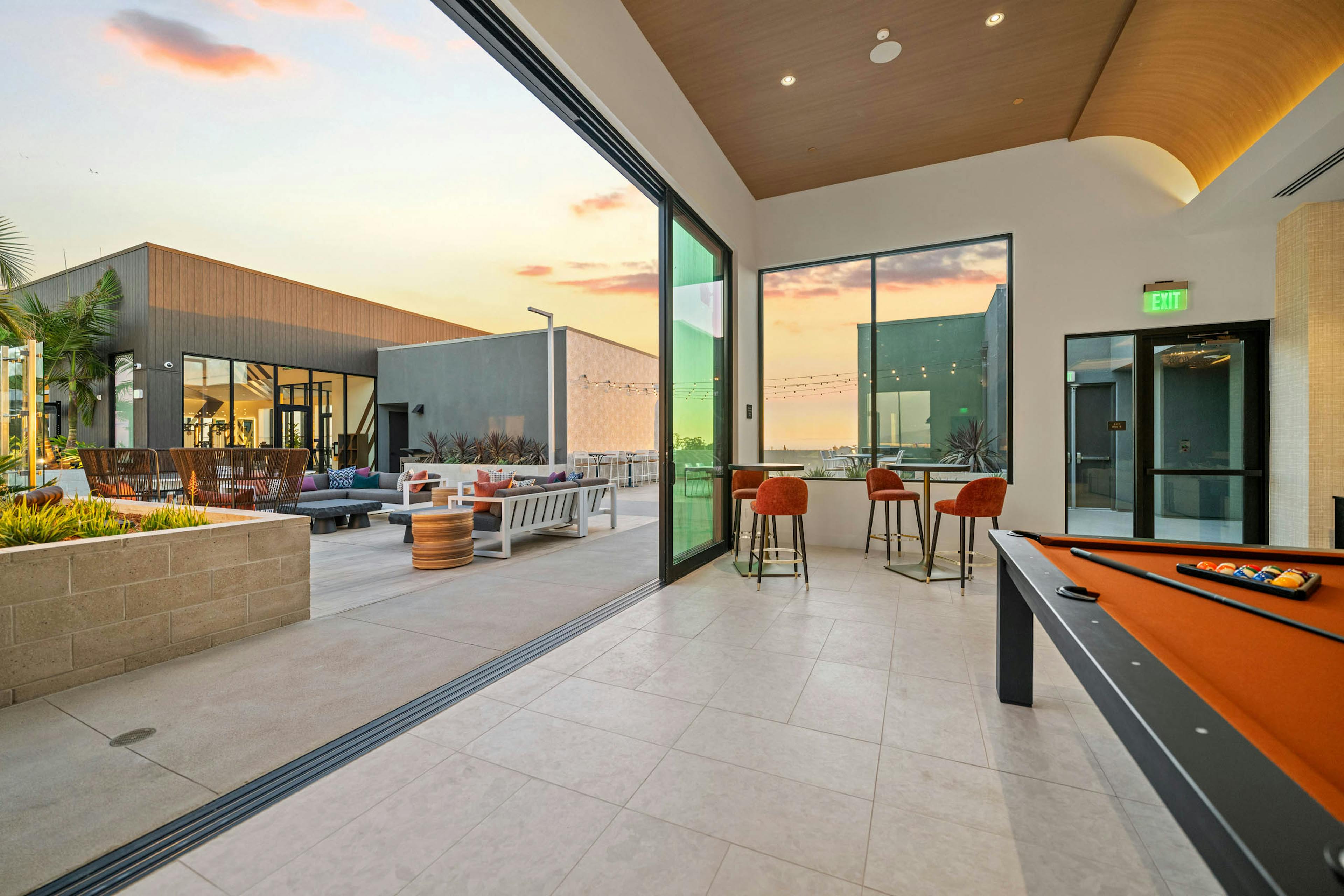 Rooftop clubroom at AMLI Aero apartments with pool table, tall windows, bar seating, and doors opening to rooftop terrace at sunset.