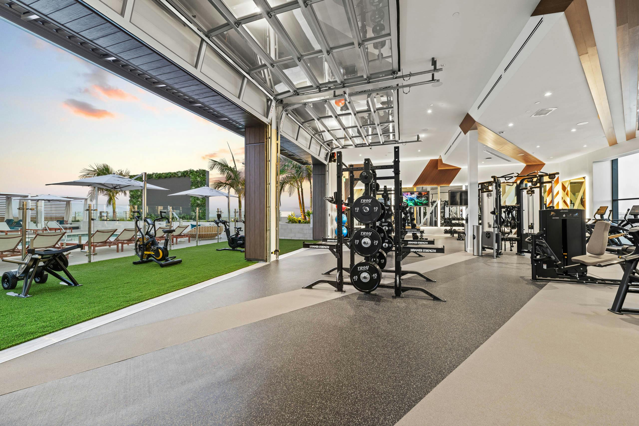 Indoor outdoor fitness at AMLI Aero apartments with weight racks inside and stationary bikes on turf overlooking rooftop pool deck.
