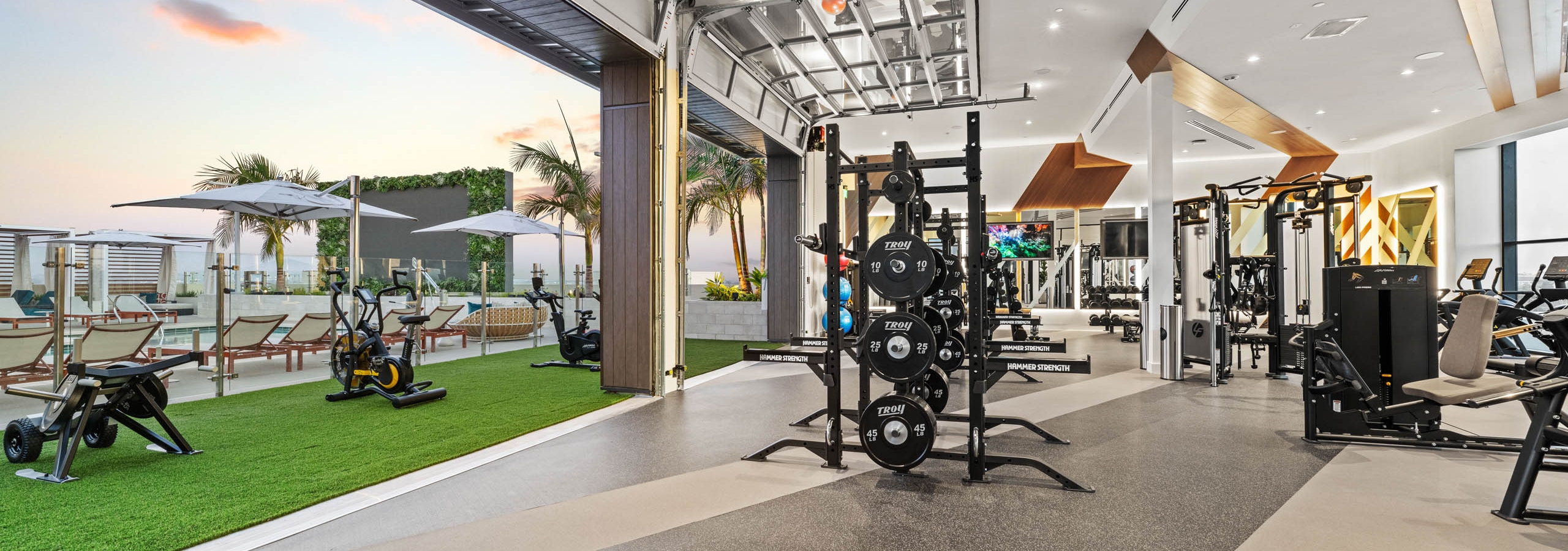 Indoor outdoor fitness at AMLI Aero apartments with weight racks inside and stationary bikes on turf overlooking rooftop pool deck.