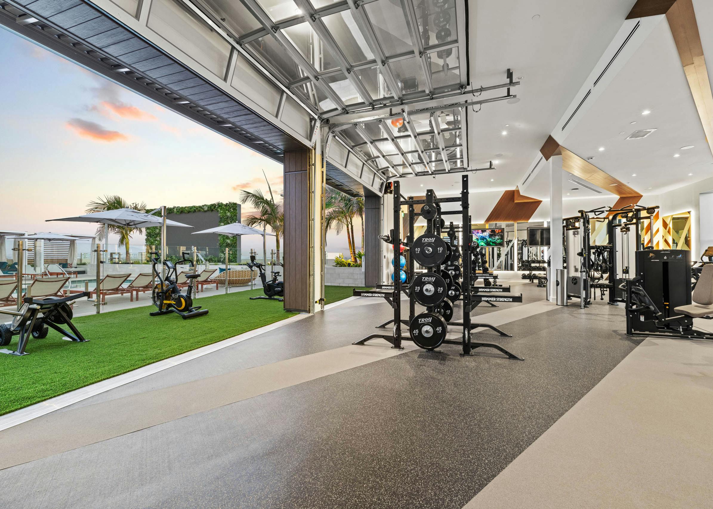 Indoor outdoor fitness at AMLI Aero apartments with weight racks inside and stationary bikes on turf overlooking rooftop pool deck.