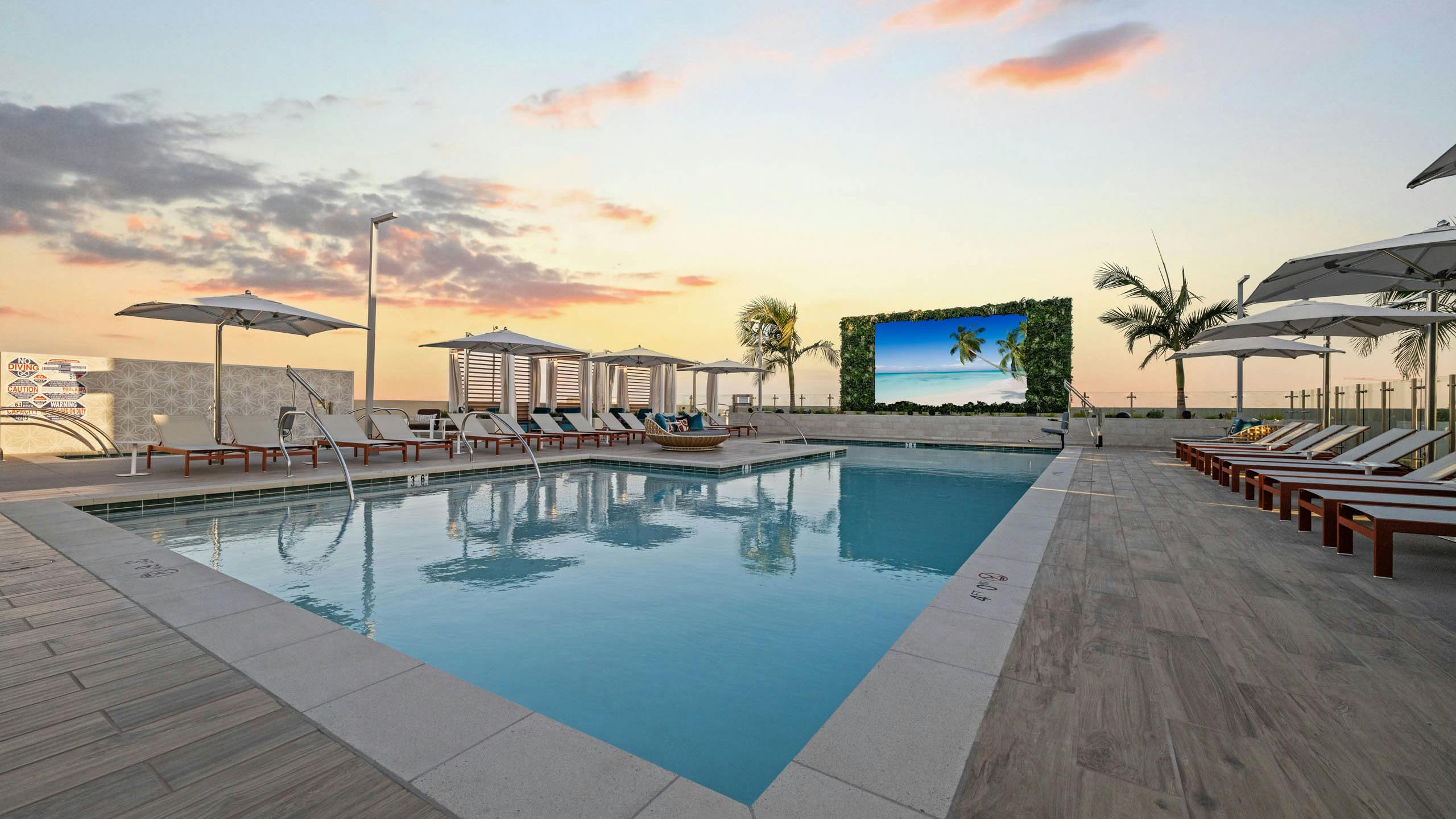Dusk view of sparkling blue pool at AMLI Aero with recliners, large tv, fitness center building and palm trees and panoramic rooftop.