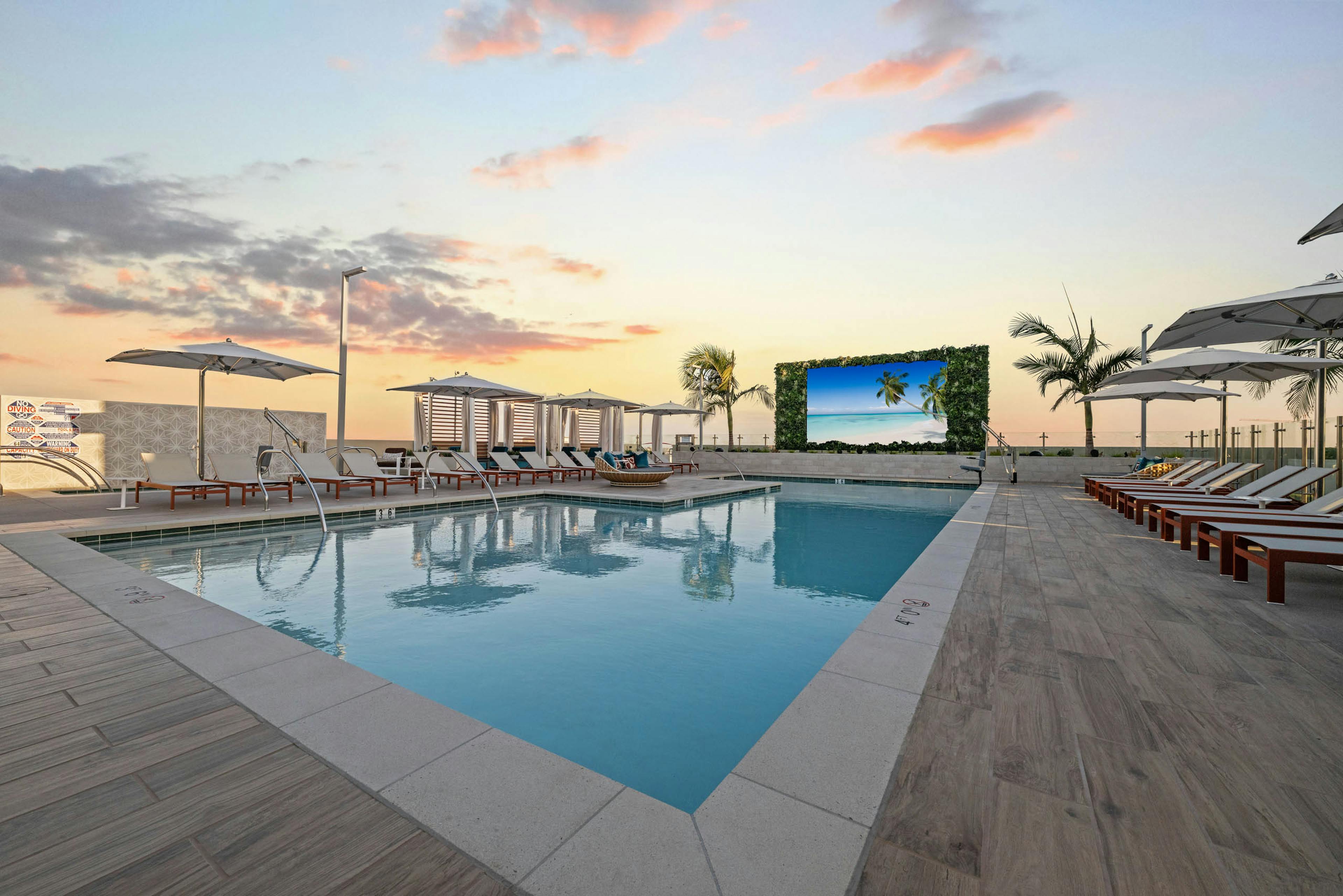Dusk view of sparkling blue pool at AMLI Aero with recliners, large tv, fitness center building and palm trees and panoramic rooftop.