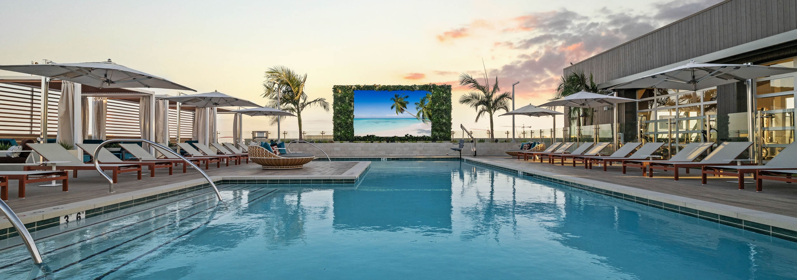 Rooftop pool deck at AMLI Aero apartments with lounge seating, cabanas, umbrellas, and large outdoor TV screen at dusk.