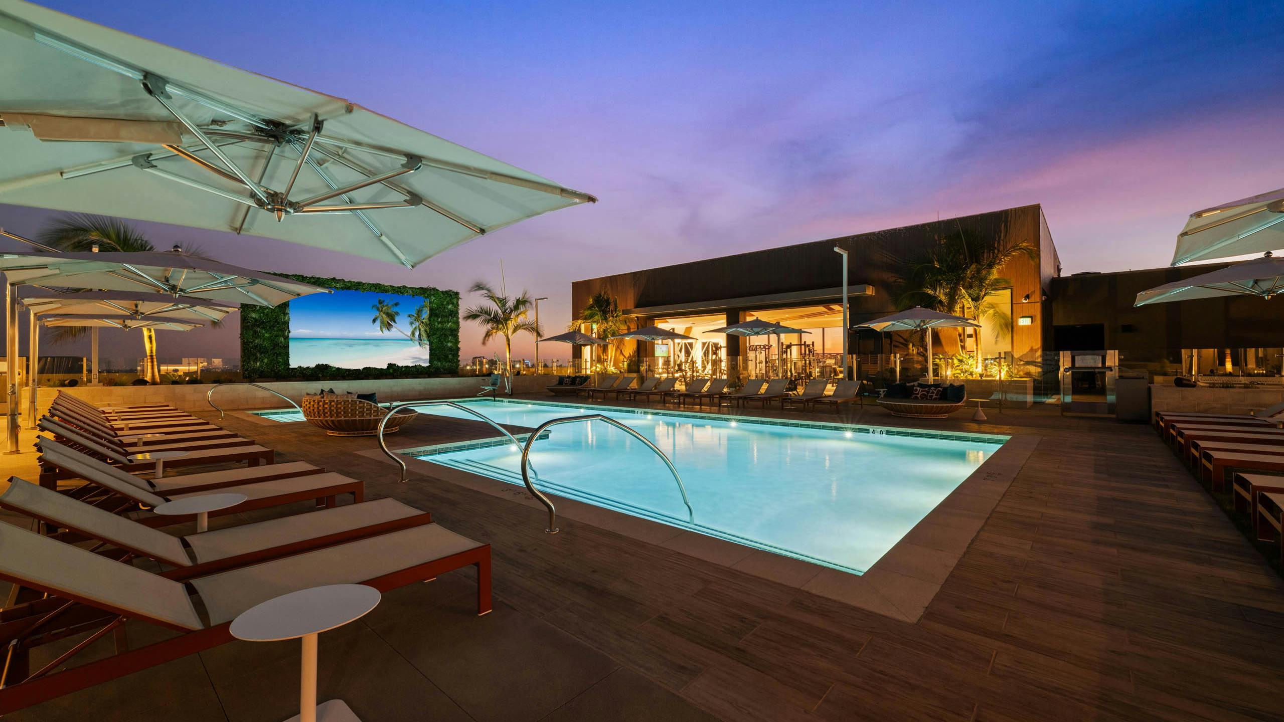 Rooftop pool deck at AMLI Aero apartments with lounge seating, cabanas, umbrellas, and large outdoor TV screen at dusk.