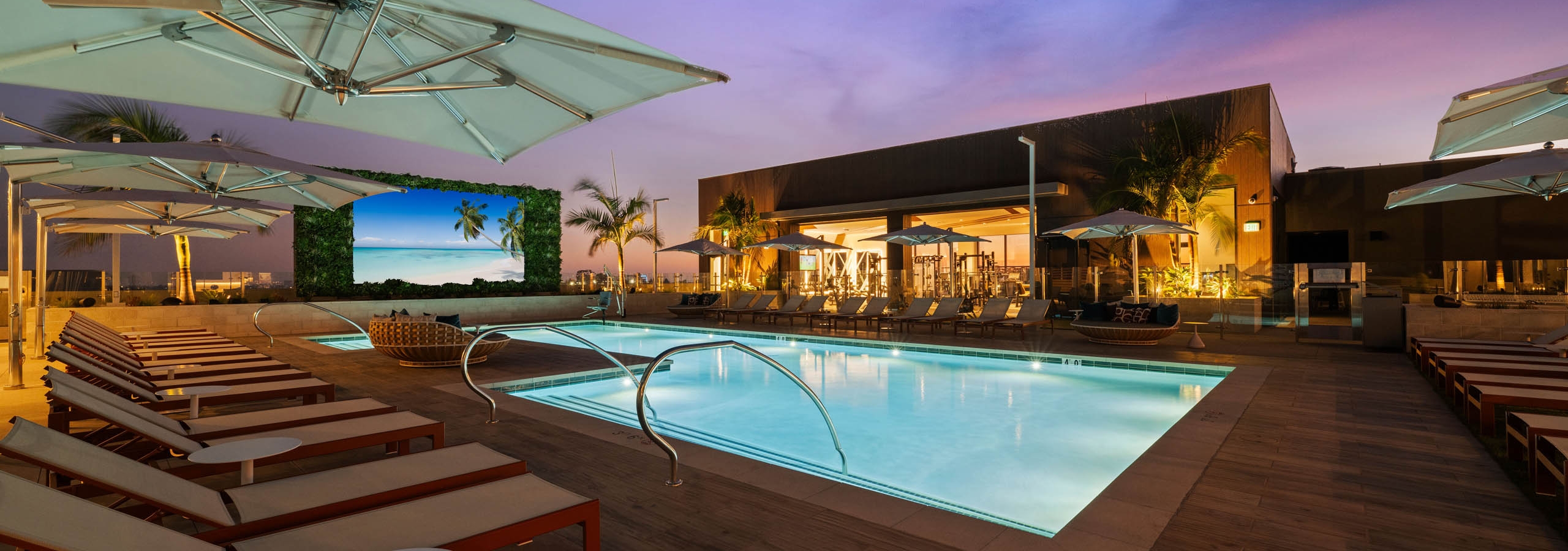 Rooftop pool deck at AMLI Aero apartments with lounge seating, cabanas, umbrellas, and large outdoor TV screen at dusk.