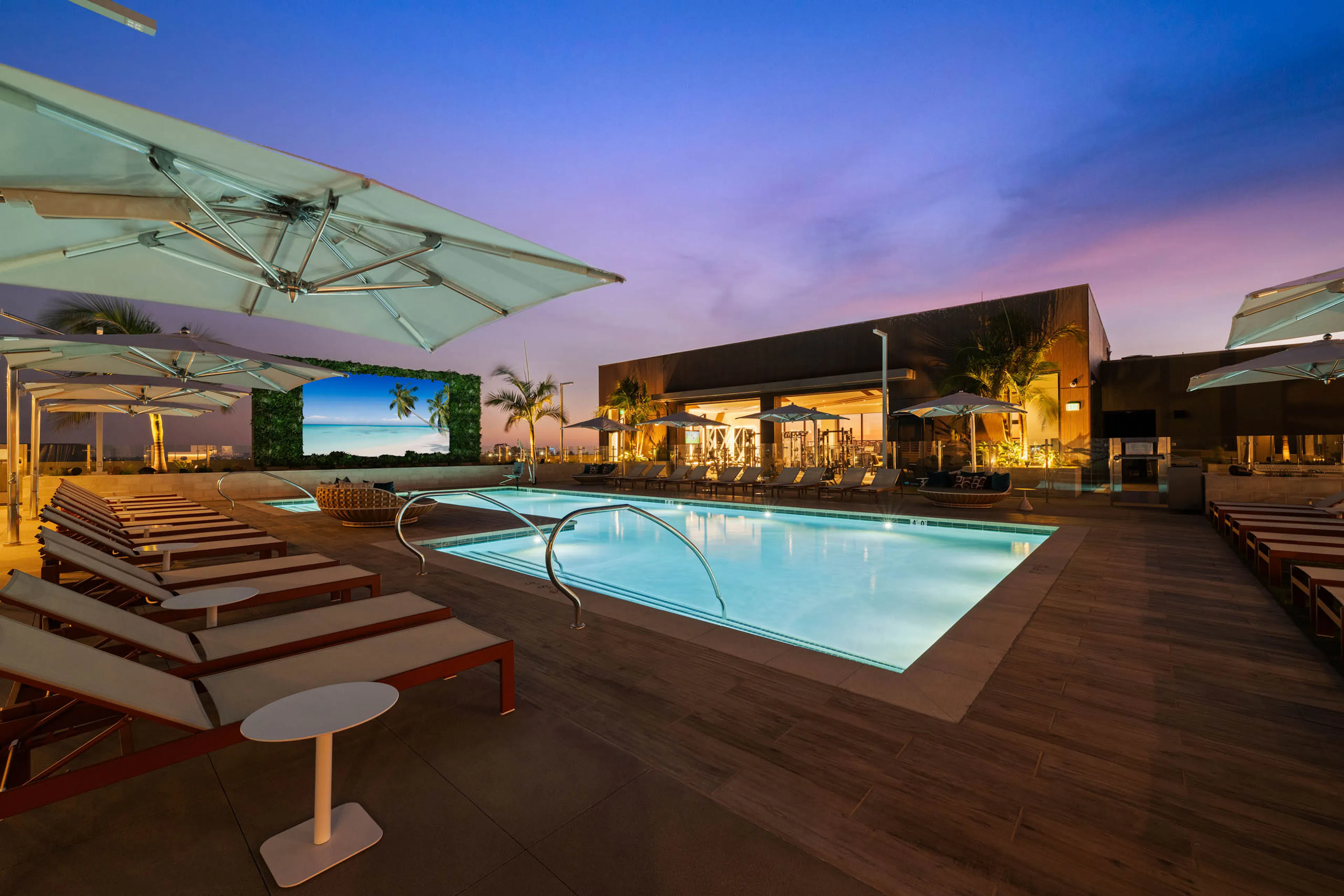 Rooftop pool deck at AMLI Aero apartments with lounge seating, cabanas, umbrellas, and large outdoor TV screen at dusk.