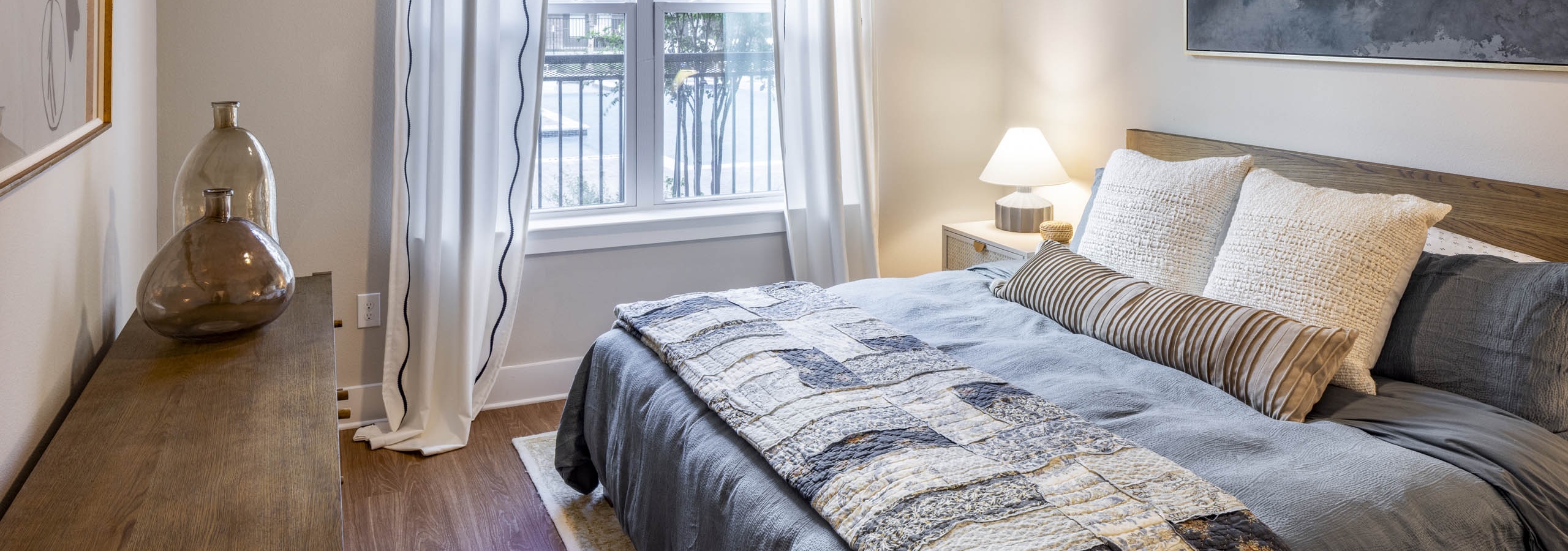 Bedroom at AMLI North Park apartments with gray bedding pillows large window with white curtains nightstand lamp and wood dresser