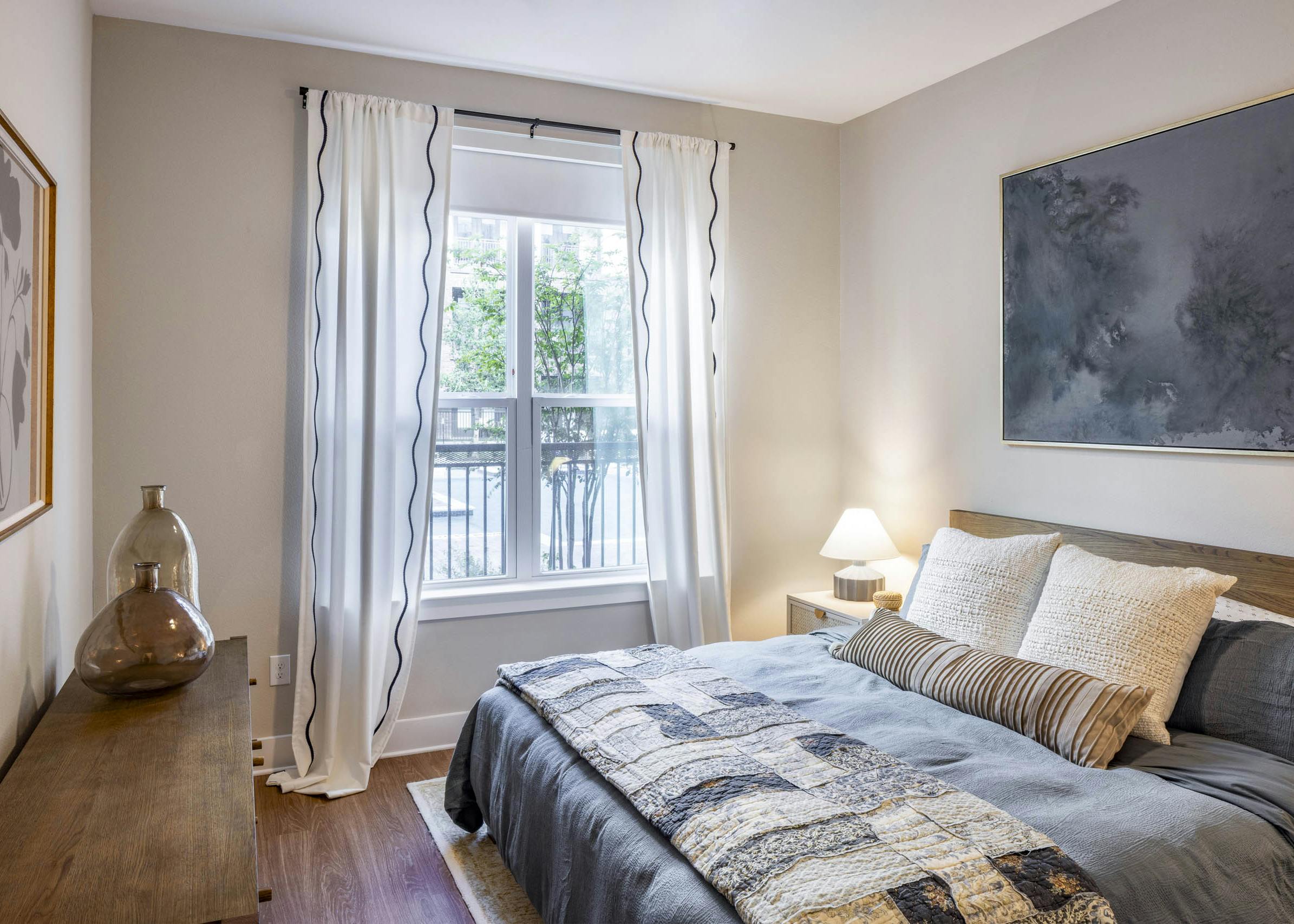 Bedroom at AMLI North Park apartments with gray bedding pillows large window with white curtains nightstand lamp and wood dresser