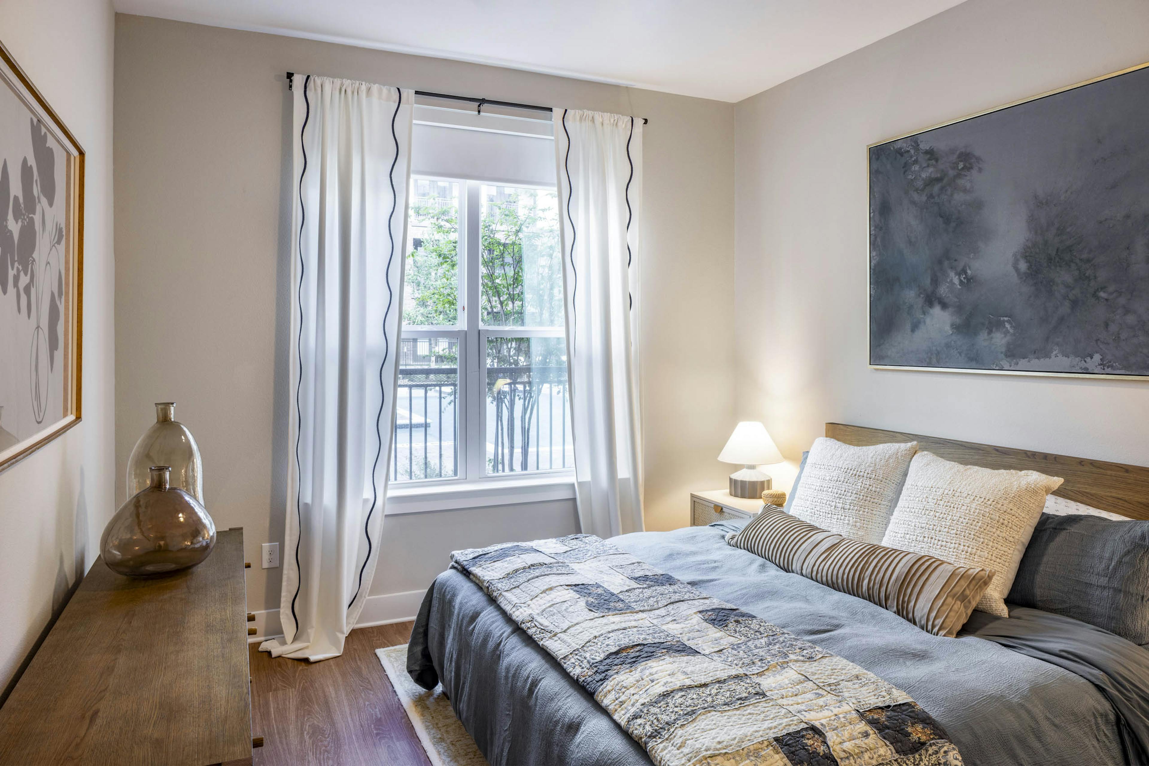 Bedroom at AMLI North Park apartments with gray bedding pillows large window with white curtains nightstand lamp and wood dresser