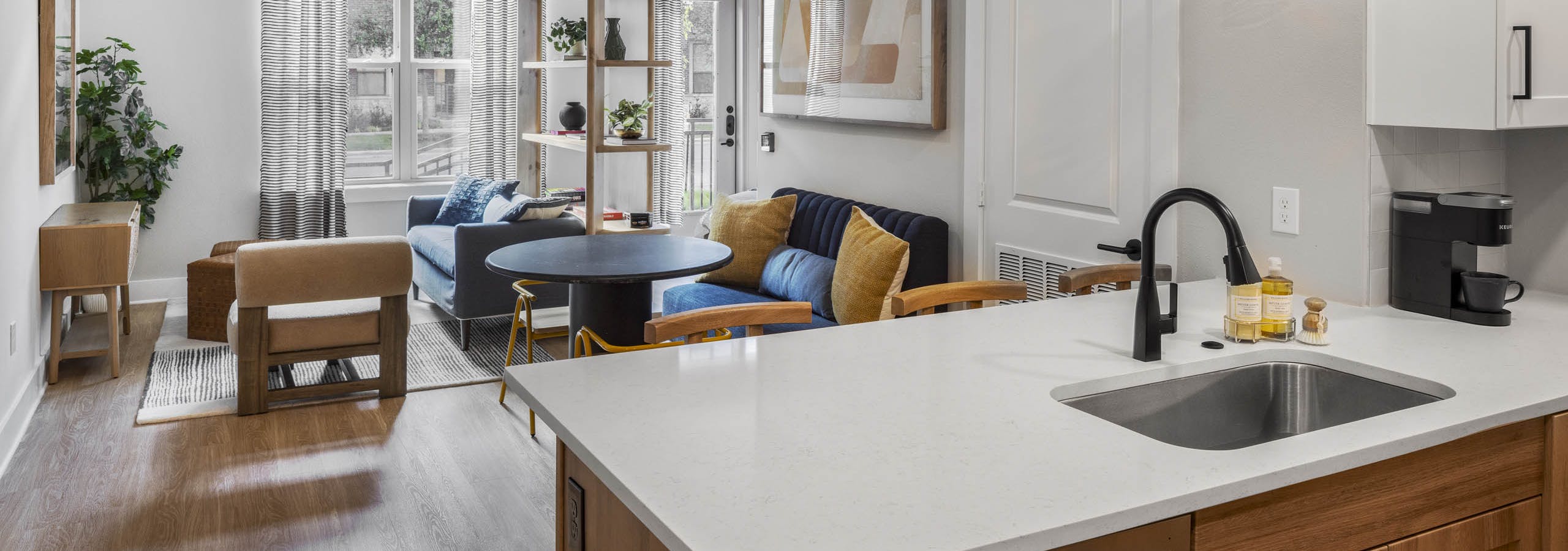 Open concept AMLI North Park apartment interior with kitchen white countertop pendant lights blue sofa yellow pillows and round table