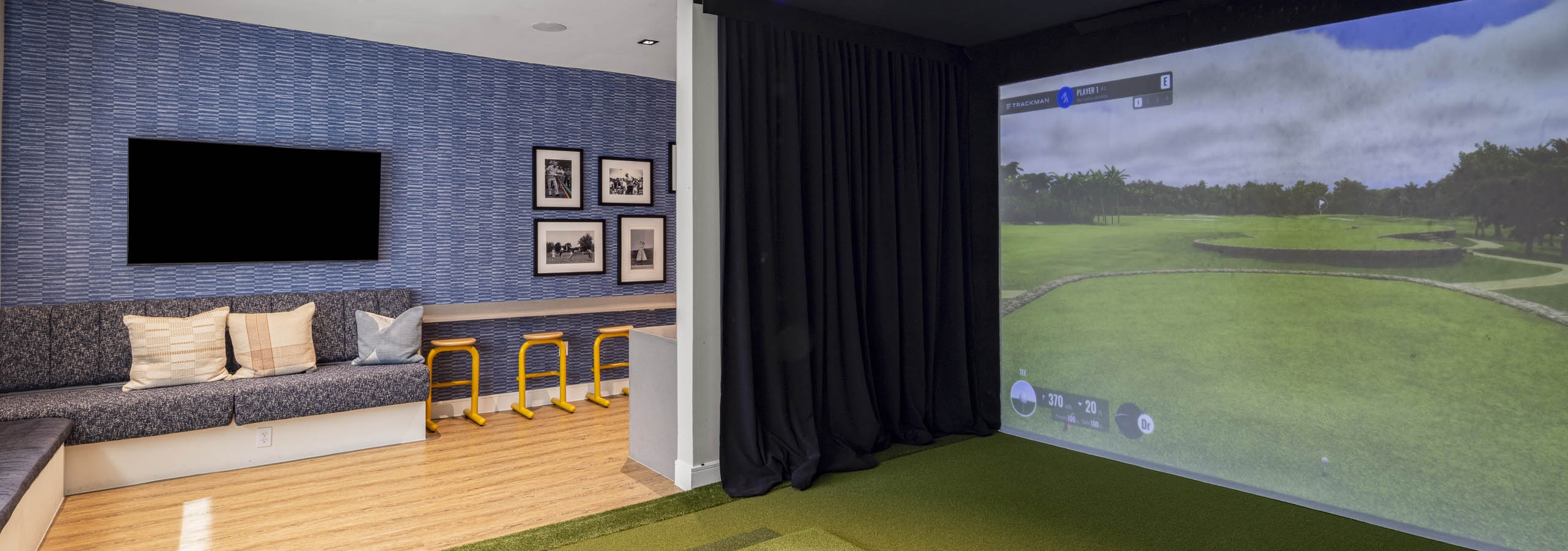 Indoor golf simulator room at AMLI North Park apartment community with screen turf mat bench seating yellow stools and wall décor