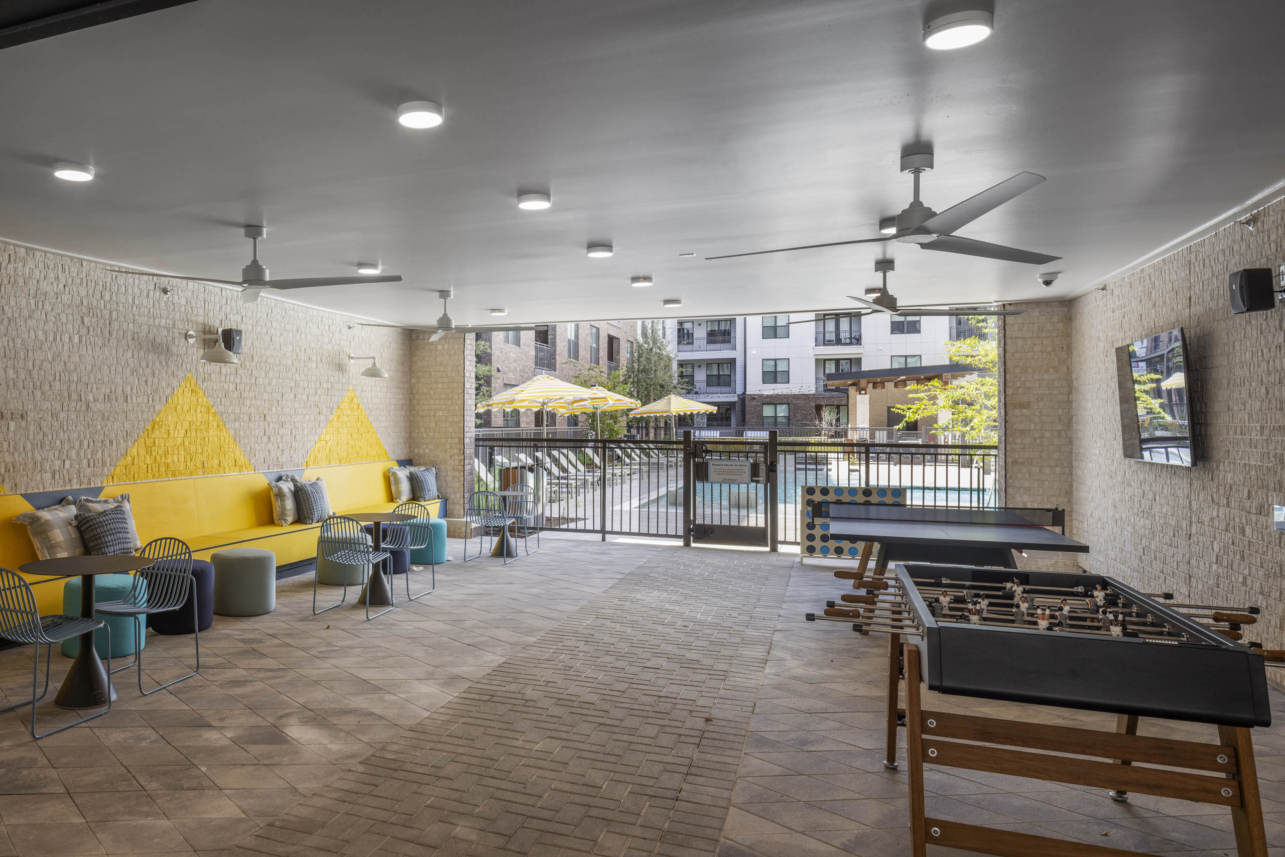 Covered patio at AMLI North Park apartment community with yellow cushioned benches round tables foosball tables TV and view of pool area