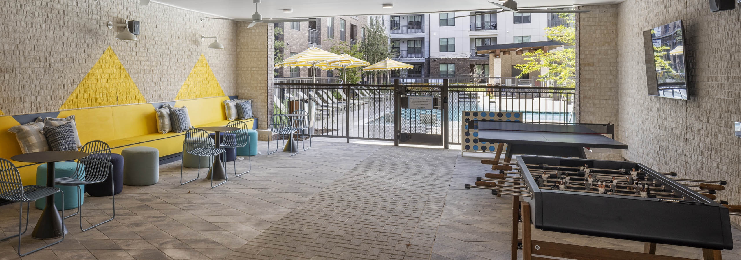 Covered patio at AMLI North Park apartment community with yellow cushioned benches round tables foosball tables TV and view of pool area