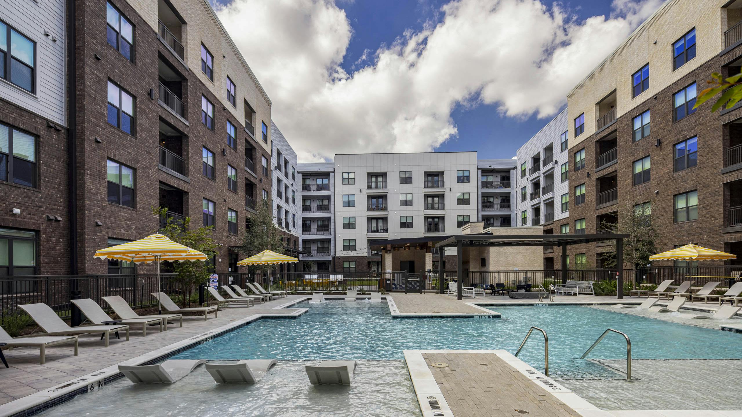 Outdoor swimming pool at AMLI North Park apartment community with submerged lounge chairs yellow umbrellas brick and lush landscaping