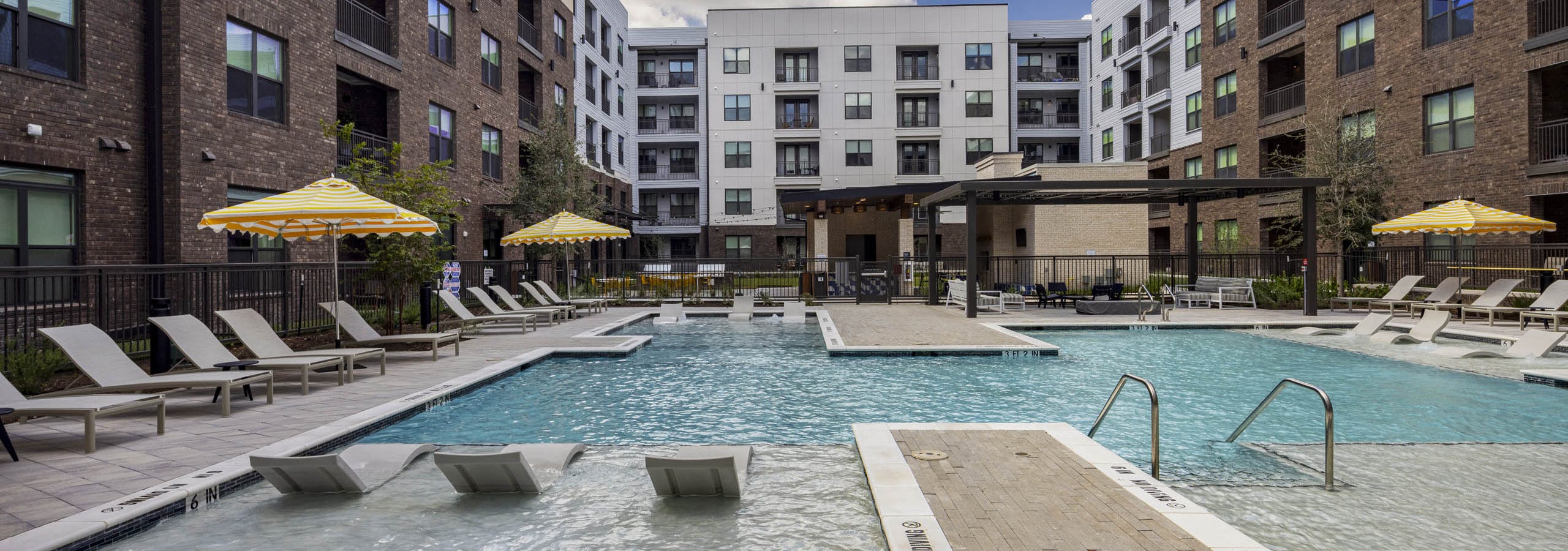 Outdoor swimming pool at AMLI North Park apartment community with submerged lounge chairs yellow umbrellas brick and lush landscaping