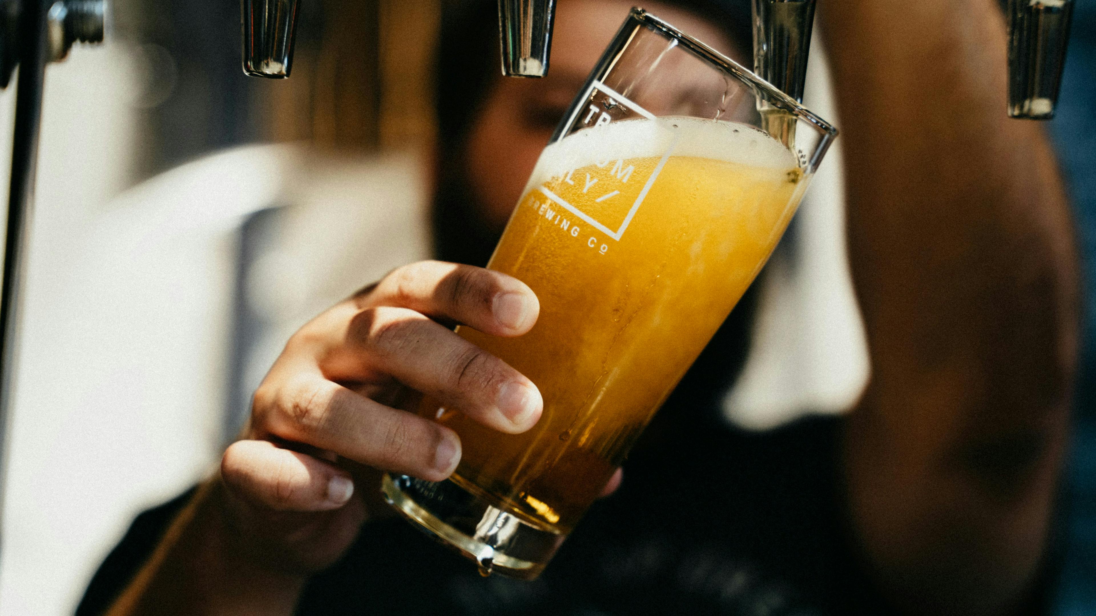 A close-up, high-angle shot of a person filling a large beer glass from a shiny metal beer tap. The glass is tilted at an angle.
