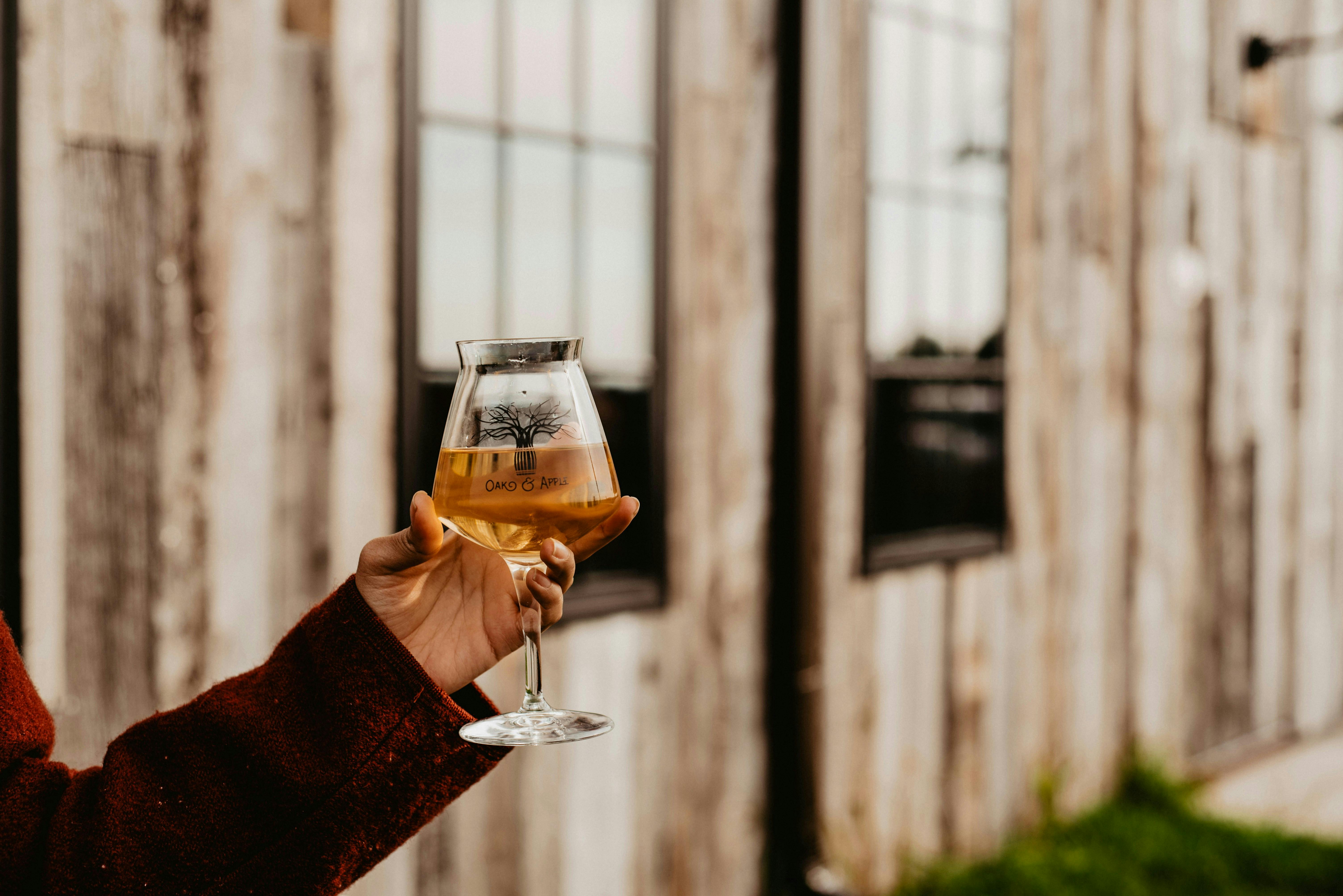 A person's hand holds up a stemmed glass filled with a light-colored beer, with a rustic, wood-paneled building with two windows in the blurred background.
