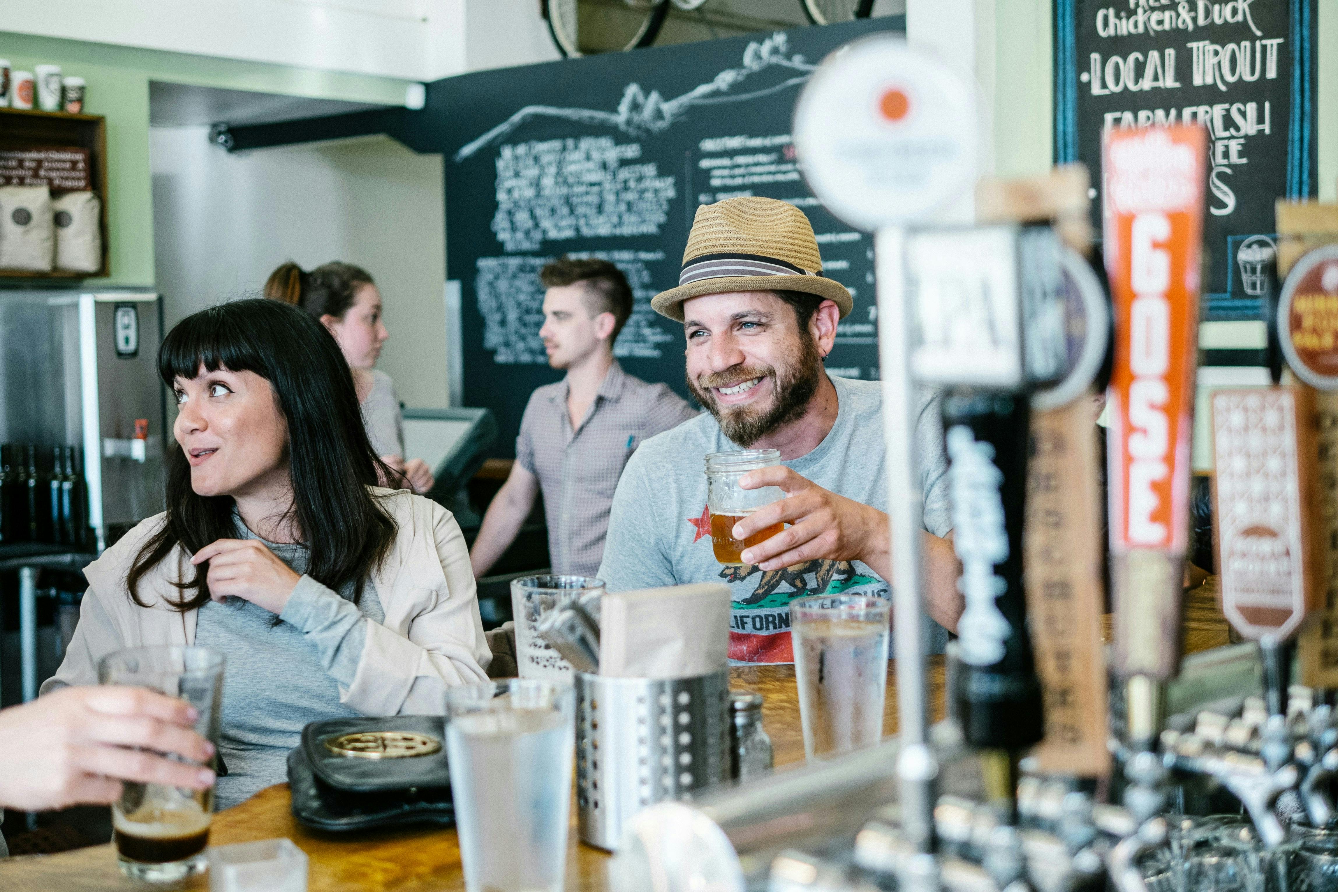 A person with a beard and a straw hat is sitting at a bar, smiling and holding a drink in a glass jar. A woman with dark hair is sitting next to him.