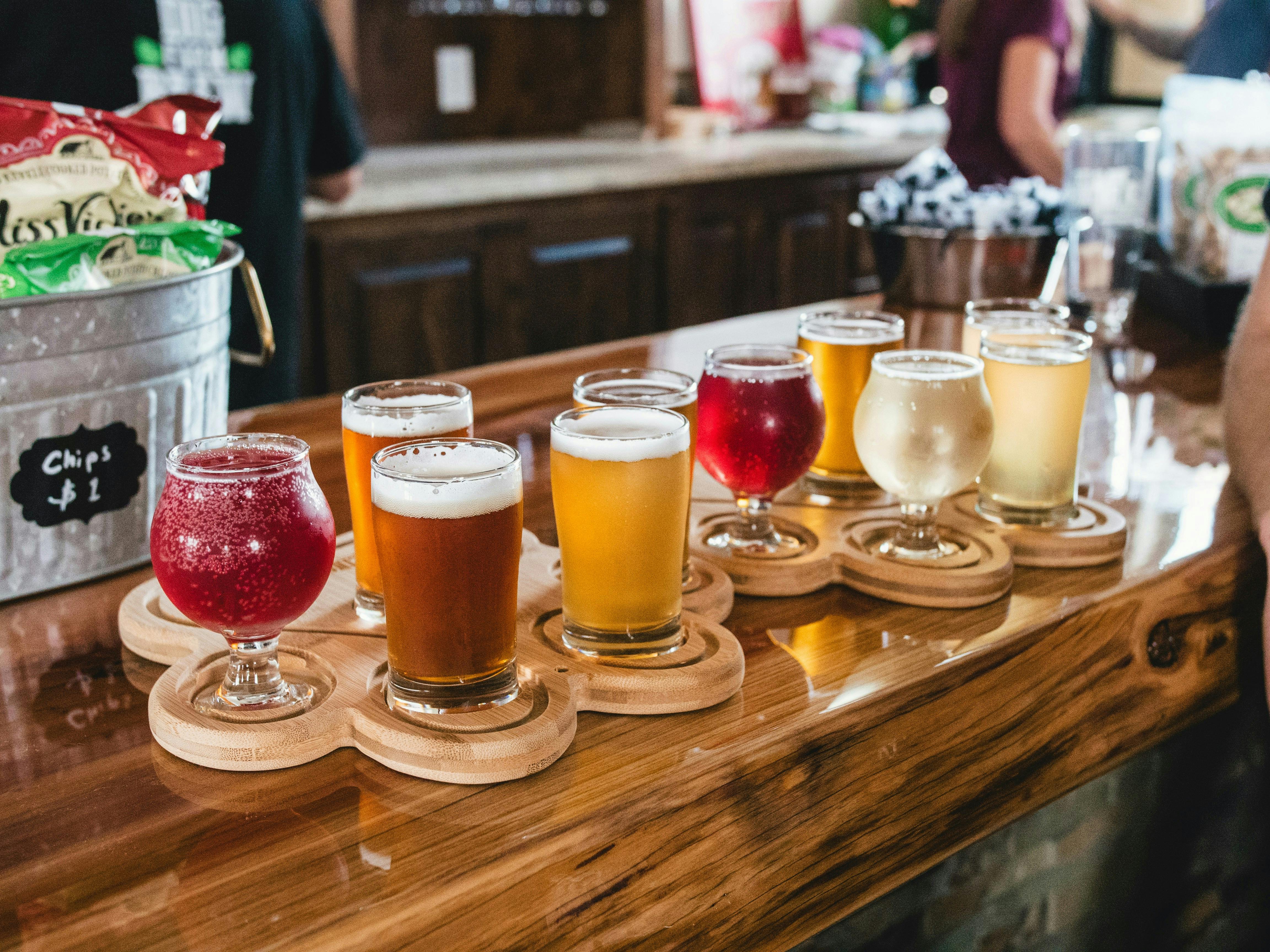 An array of beer flights, each with a different color of beer, are arranged on a wooden bar top. A bucket of chips is visible in the background.