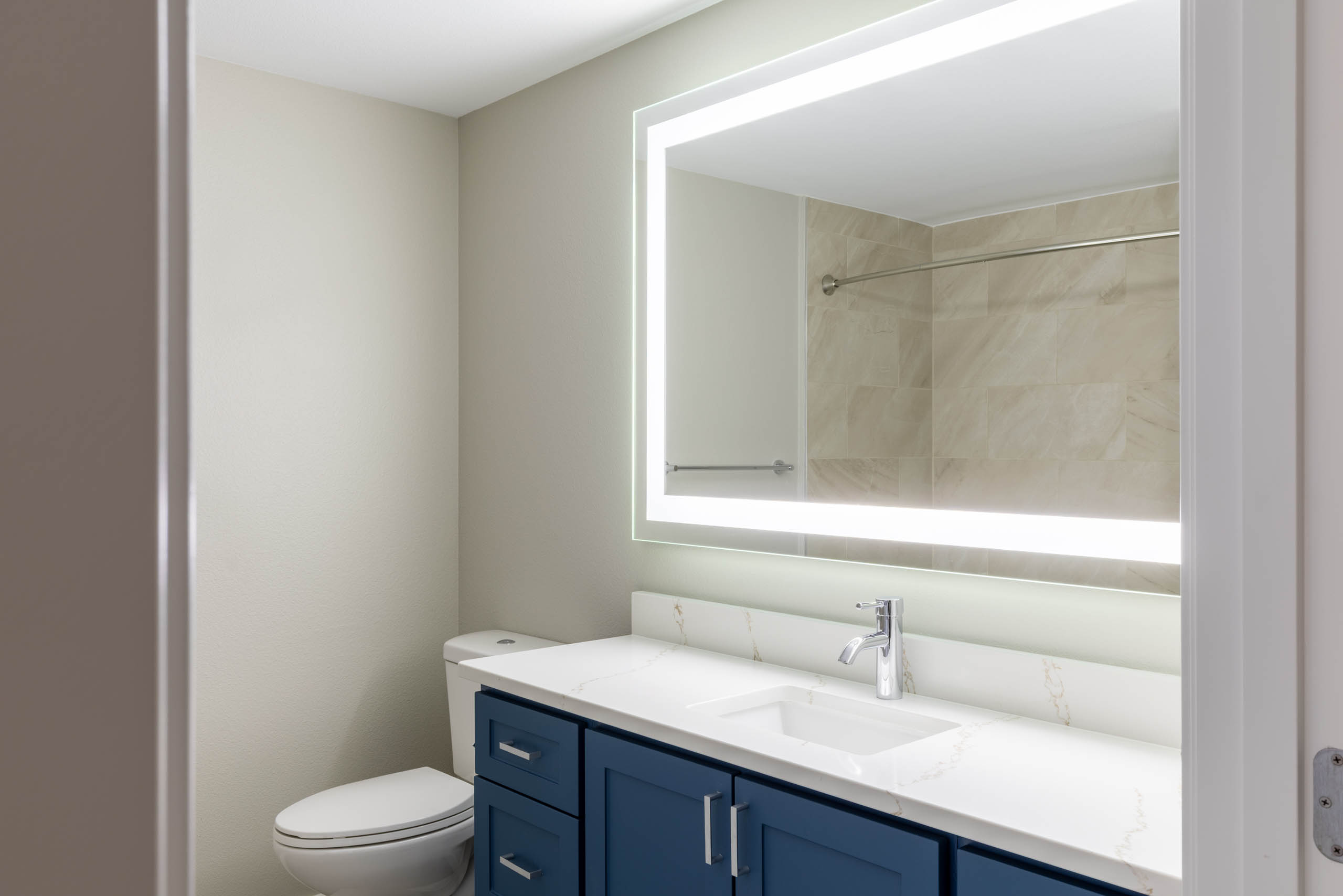 Interior view of apartment bathroom with blue vanity cabinets, large lighted mirror, and tiled shower surround at AMLI 535.