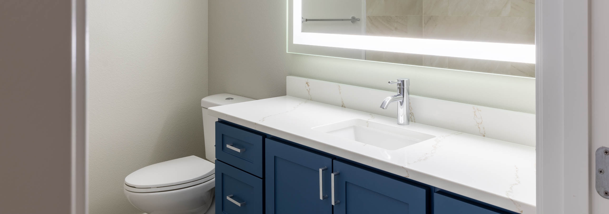 Interior view of apartment bathroom with blue vanity cabinets, large lighted mirror, and tiled shower surround at AMLI 535.