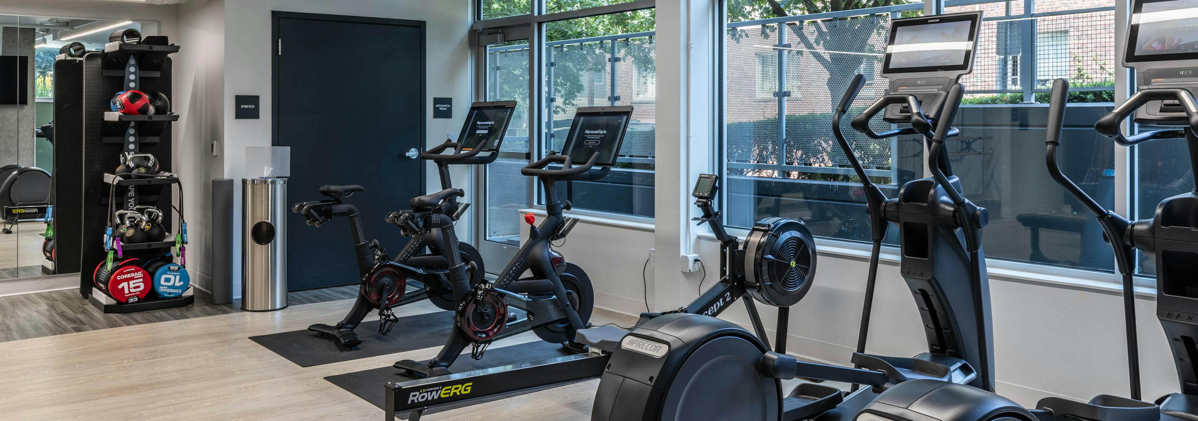 Corner view of AMLI 535 apartments fitness space with bikes, rowing machine, ellipticals, and windows overlooking trees and sidewalk.