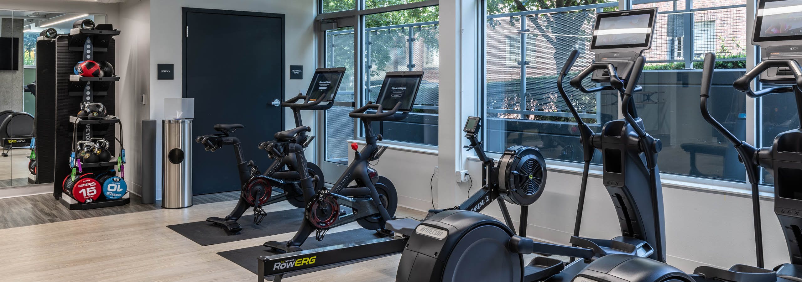 Corner view of AMLI 535 apartments fitness space with bikes, rowing machine, ellipticals, and windows overlooking trees and sidewalk.