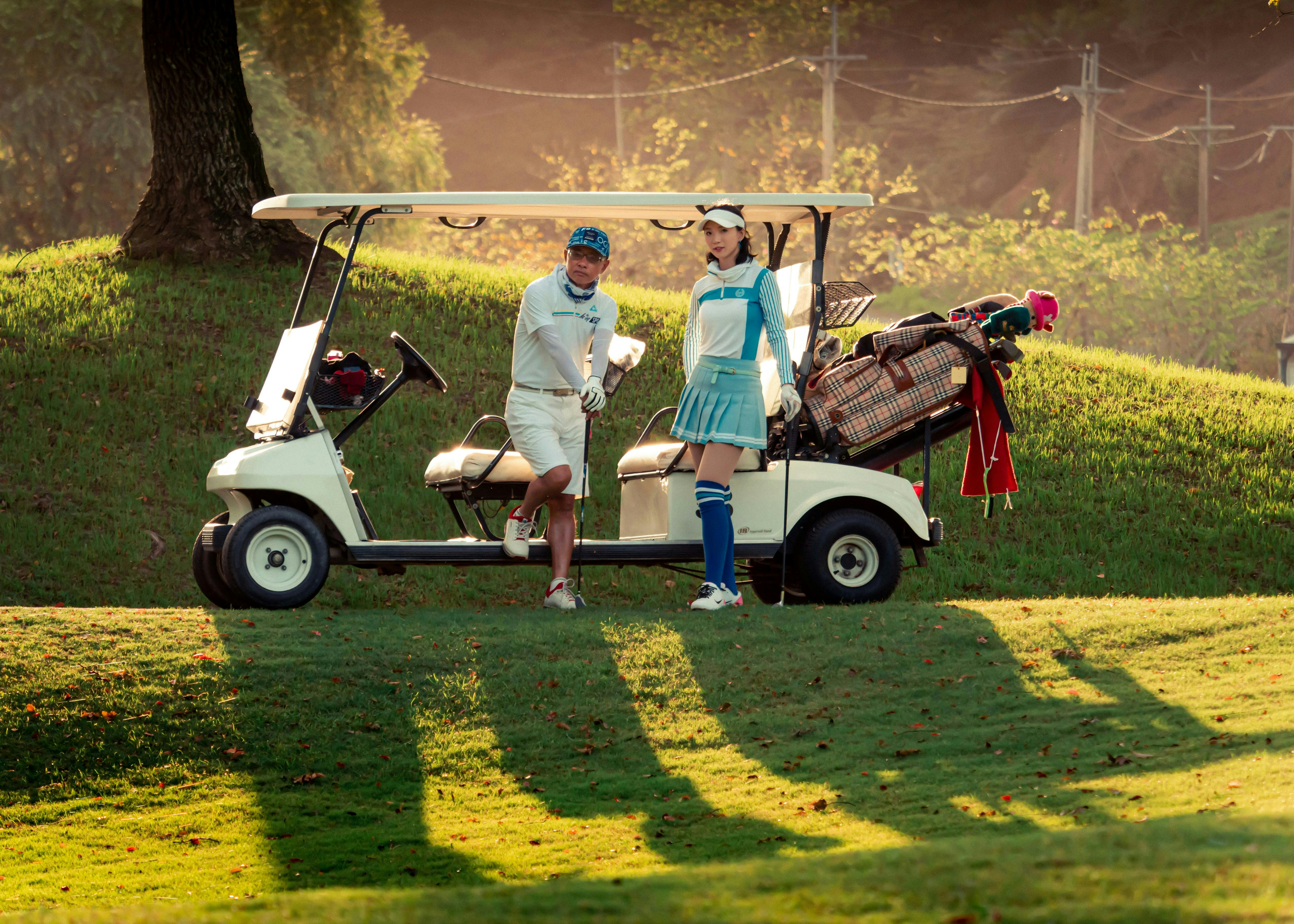 A man and a woman in golf attire stand beside a white golf cart on a grassy hill. The sun casts long shadows, and the course is lush and green. The man is smiling and the woman is looking at the camera.