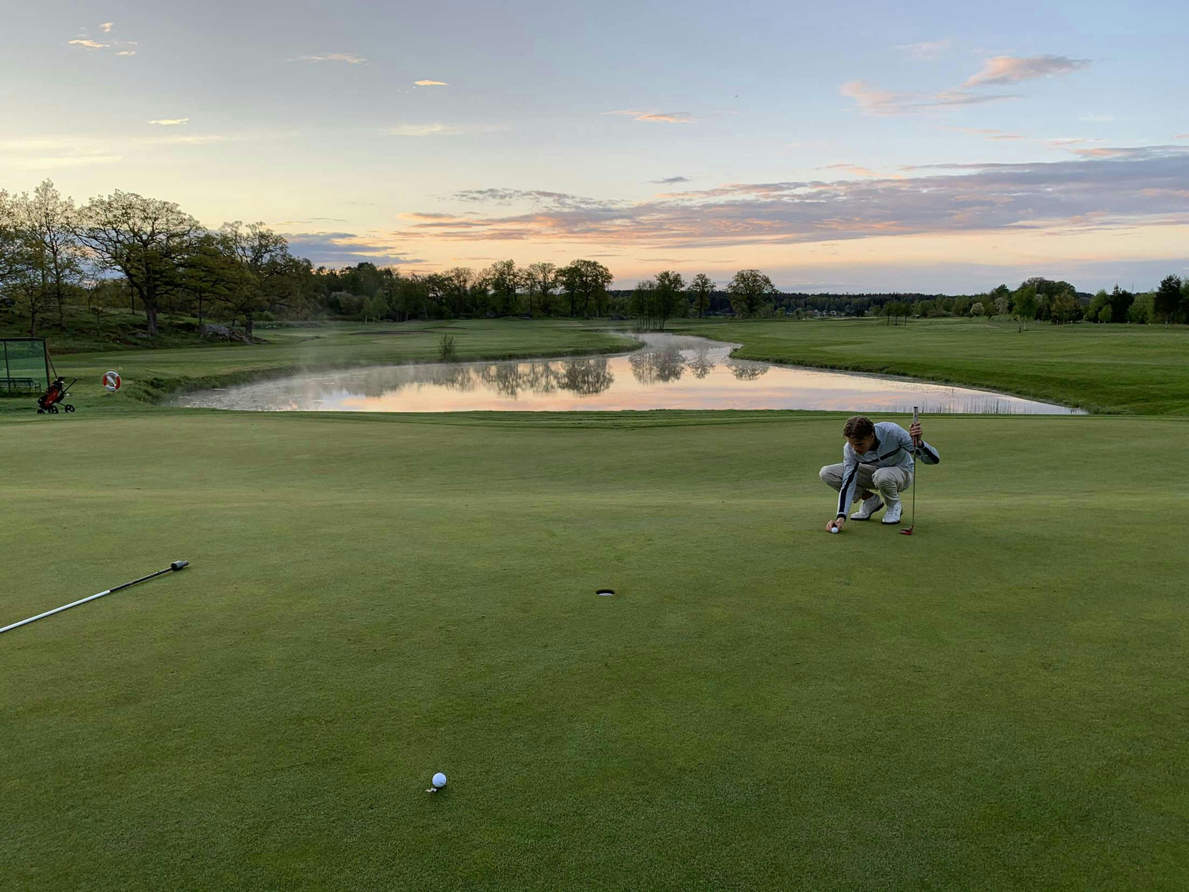 A golfer crouches on a green, manicured putting green, lining up a putt. A small pond with a slight mist sits in the background, reflecting the sunset sky. The surrounding trees are green, and the overall scene is peaceful and serene.