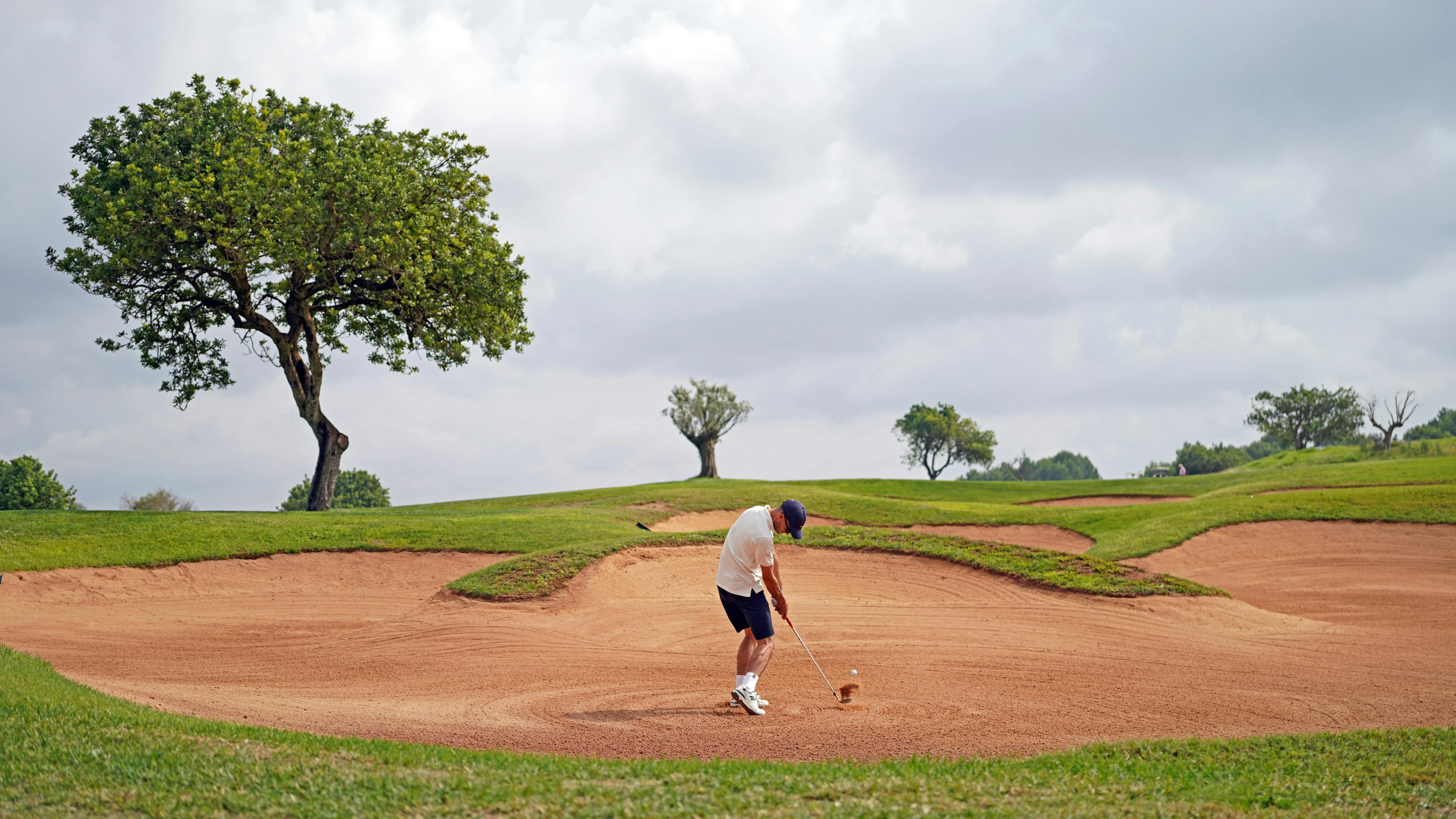 A male golfer in a white hat and shirt stands in a large sand trap, poised to hit the ball. The green grass of the course surrounds the bunker, with a large tree on the left and a few other trees in the distance. The sky is partly cloudy.